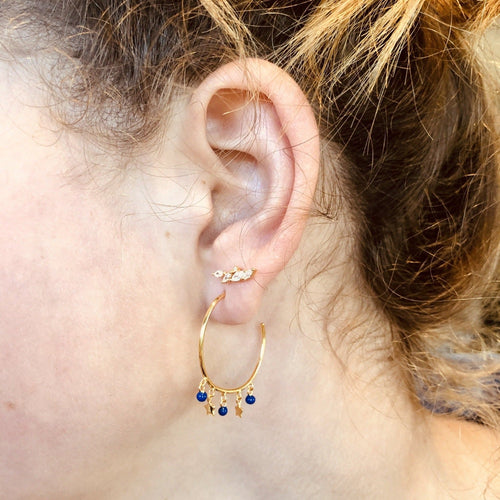 Two pairs of hoop earrings, one sterling silver and one with 18ct gold plating. Each hoop has two little star charms hanging from the bottom, with three navy enamel beads interspersed between them. Shown in a ladies ear.
