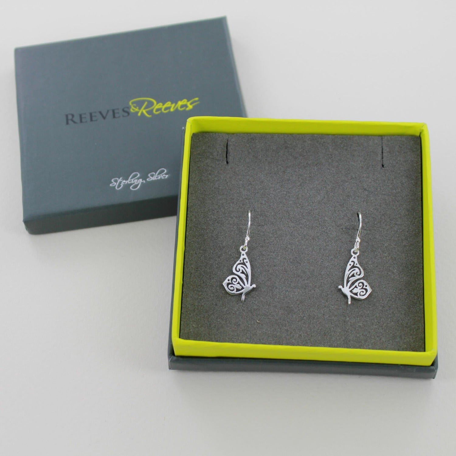 A pair of sterling silver Butterfly Drop Earrings, with cut outs in their wings and hanging on silver hooks. Shown in a Reeves & Reeves gift box.