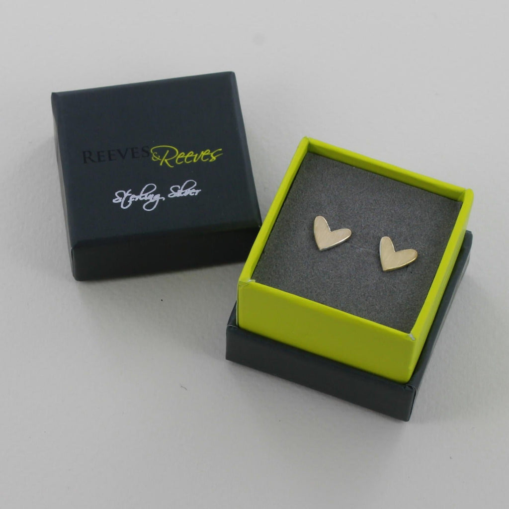 A pair of sterling silver heart stud earrings with an 18ct gold plate matt finish. Shown in a Reeves & Reeves grey and green gift box.