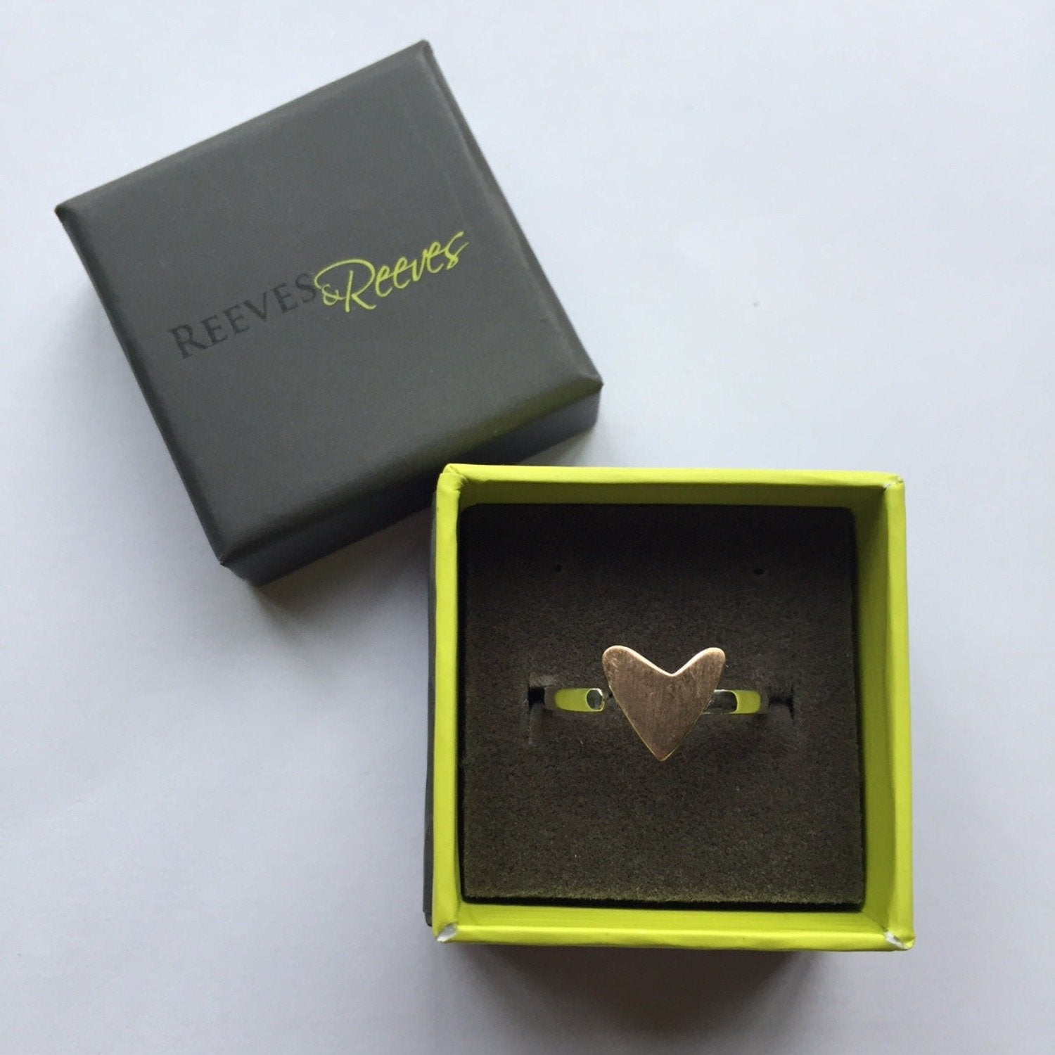 A silver adjustable banded ring with a rose gold heart as the central feature, and shown in a grey and green Reeves & Reeves branded gift box.