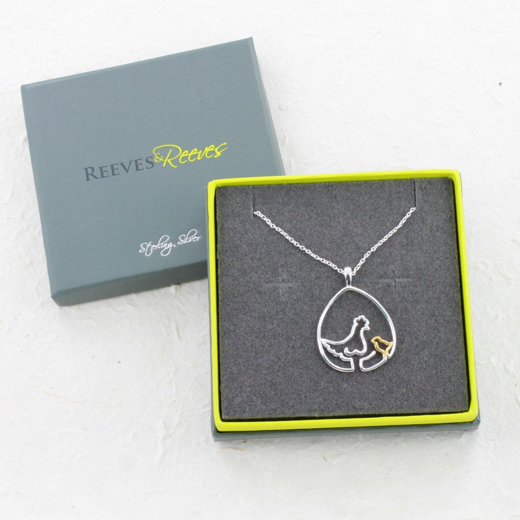 A Sterling Silver chicken pendant in a continuous line style, with a gold plated chick sat inside an egg shape pendant and hung on a silver chain. Shown in a Reeves & Reeves grey and green branded gift box.