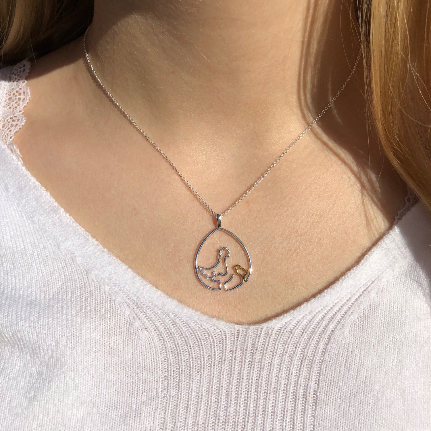 A Sterling Silver continuous line style necklace. A silver chicken starts the line in the middle, with the line continuing around the outside to create an egg shape with a gold plated chick sat inside too. Shown on a silver chain and worn on a model.