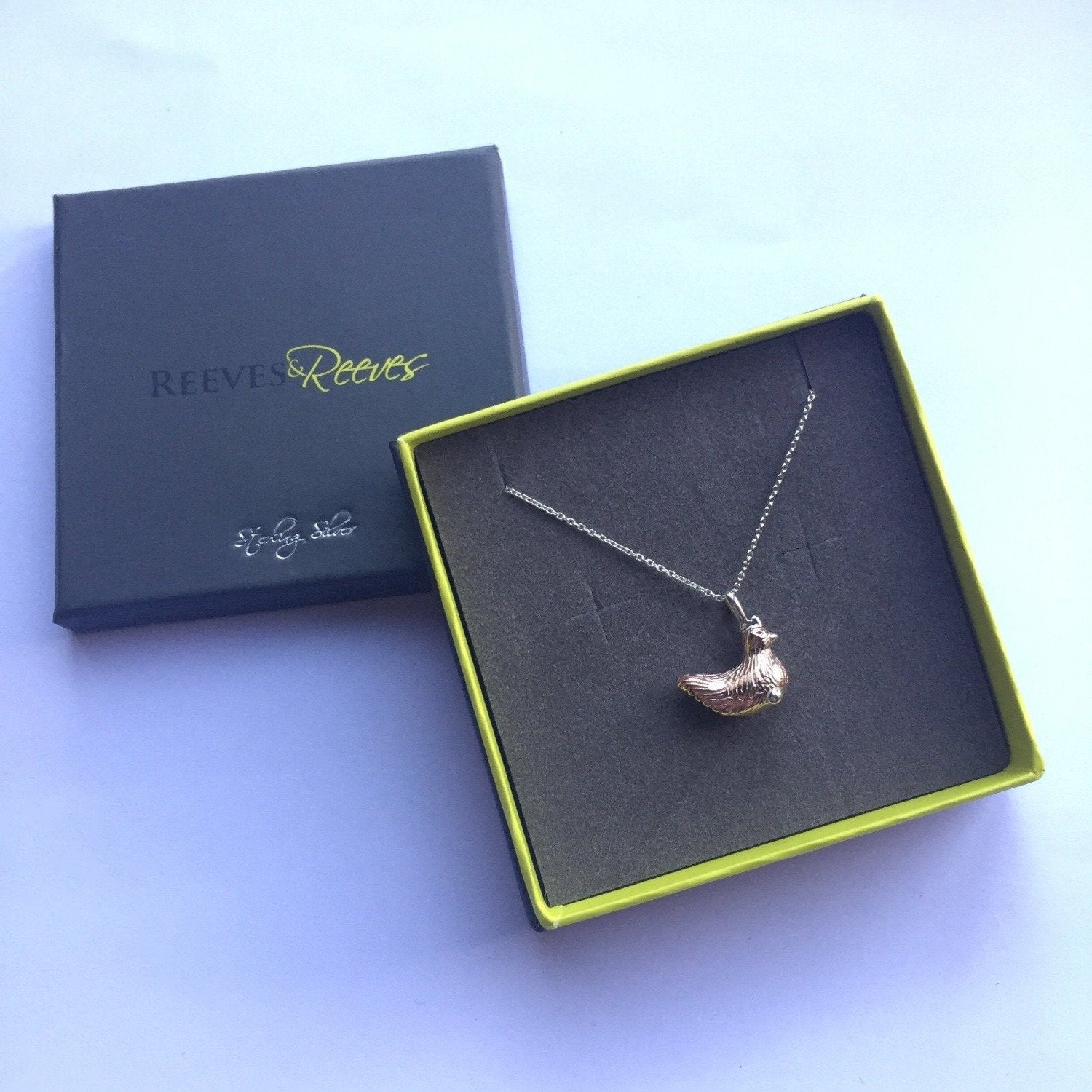 A Silver and rose gold plated Chicken Necklace, hung on a silver chain and worn in a Reeves & Reeves grey and green branded gift box.