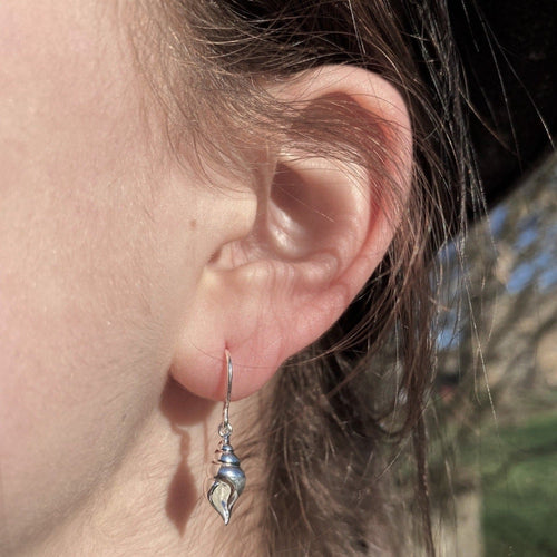 A sterling silver drop style earring with a conch shell charm hanging from the bottom of a silver hook and worn in a ladies ear.