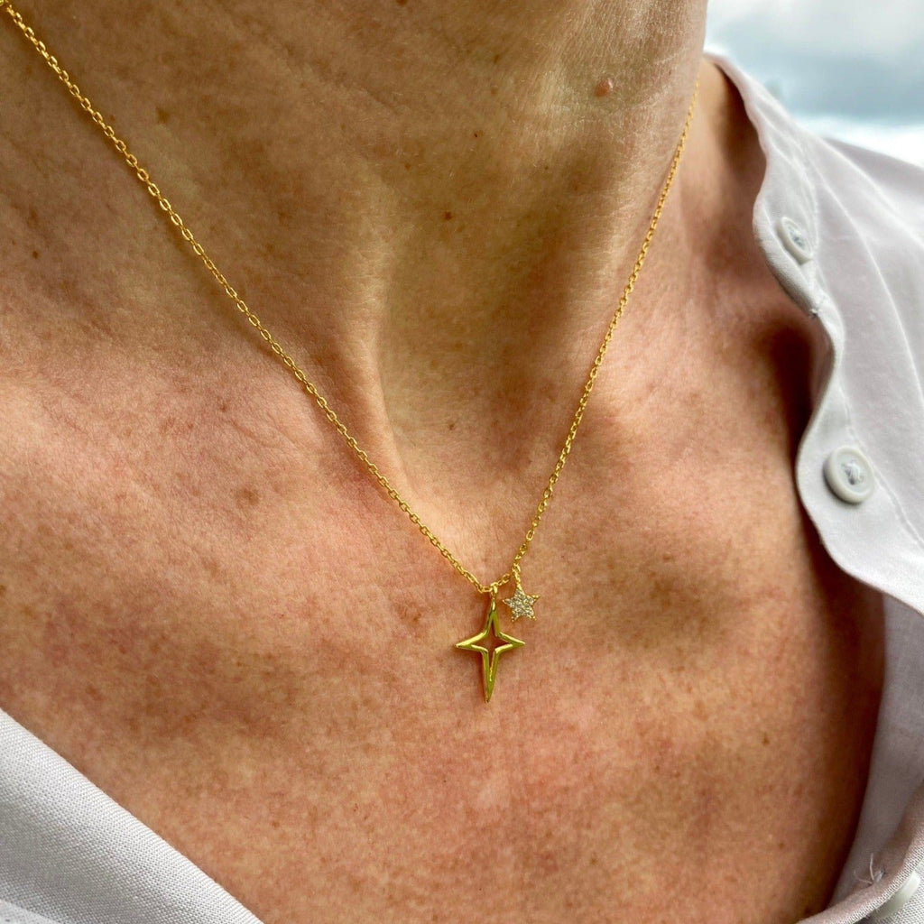 Gold necklace with four point star silhouette and a smaller five point star encrusted with cubic zirconia stones, hanging on a fine gold plated chain, worn on a models neck.