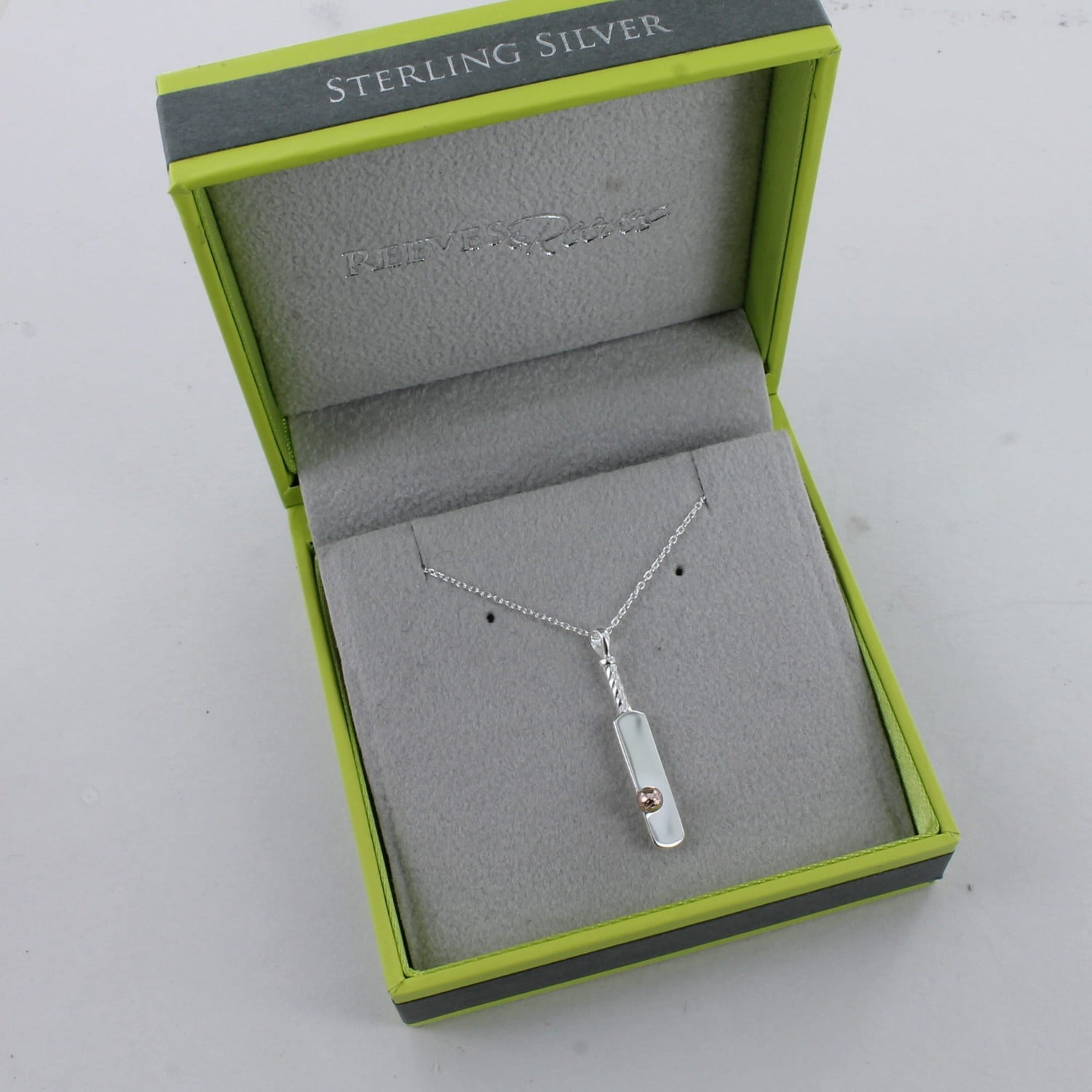 A silver cricket bat with a rose gold plated ball on the front and on a silver chain. Shown in a Reeves & Reeves branded gift box.