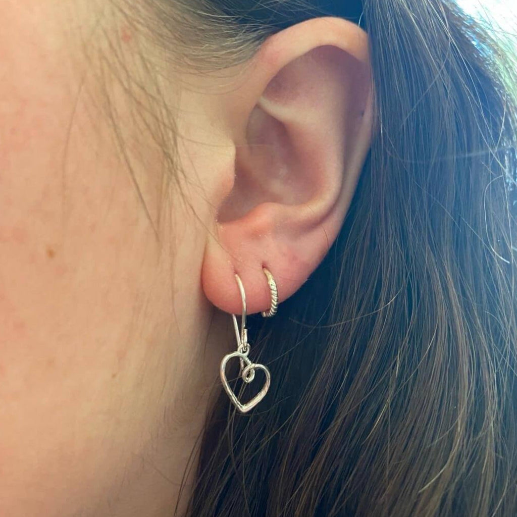 A sterling silver curly heart silhouette hanging from the bottom of a silver hook and shown in a ladies ear.