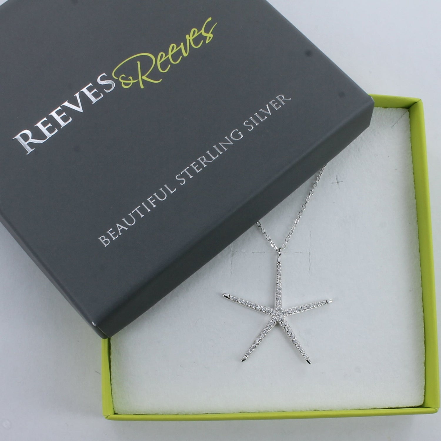 A Silver Sparkling Starfish Necklace, filled with cubic zirconia stones hanging on a silver chain and shown in a Reeves & Reeves grey and green branded gift box.