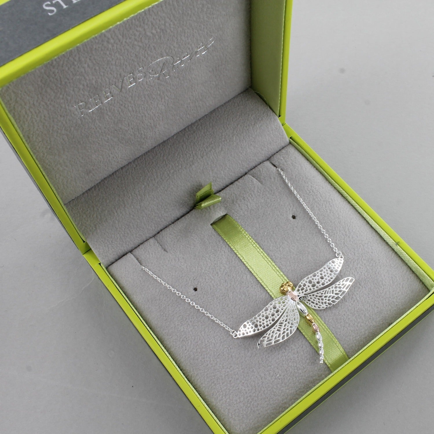 A Sterling Silver Dragonfly Necklace, with cut out wings and 18ct yellow and rose gold plate finish on its body. Hung on a silver chain and shown in a Reeves & Reeves grey and green branded box.