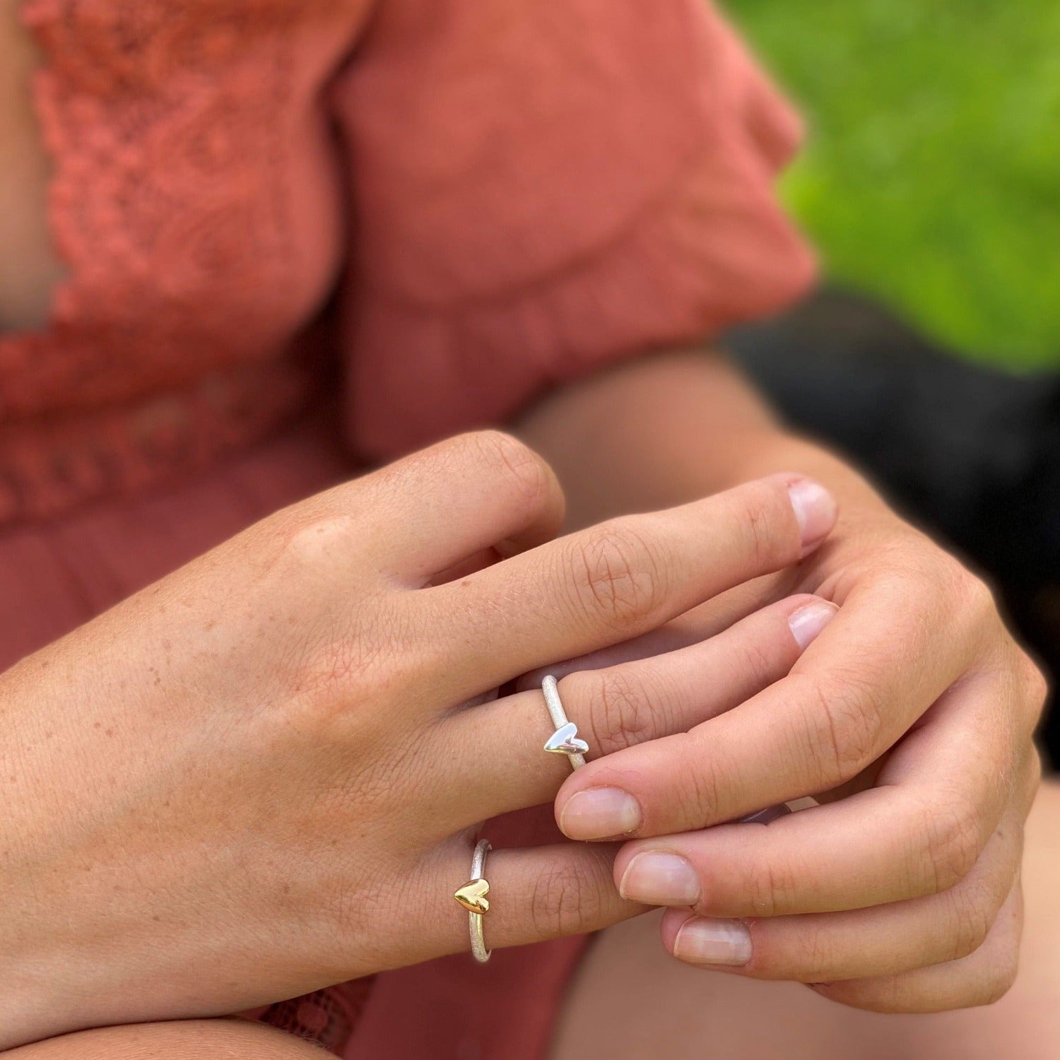 Two silver rings worn on a ladies fingers, one is silver with a silver heart and the other a silver band with an 818ct gold plated heart.
