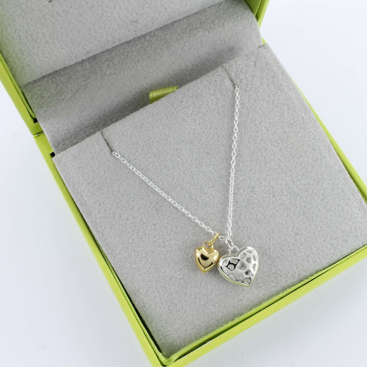 A Sterling Silver Heart with cut outs on the front and a smaller solid gold heart sat next to it on the chain. Shown in a grey and green gift box.