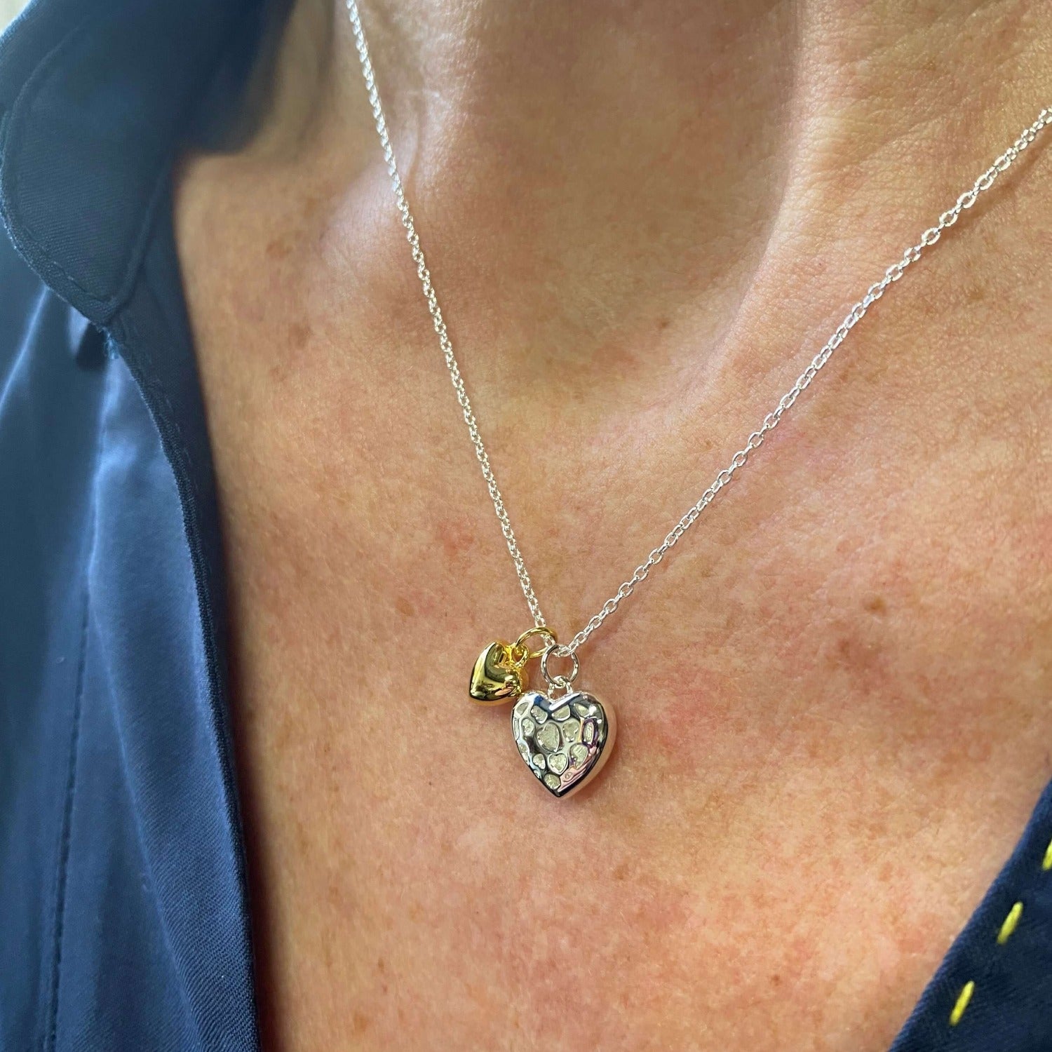 A Sterling Silver Heart with cut outs on the front, sat next to a solid gold plated heart charm, hung on a silver chain and worn on a model.