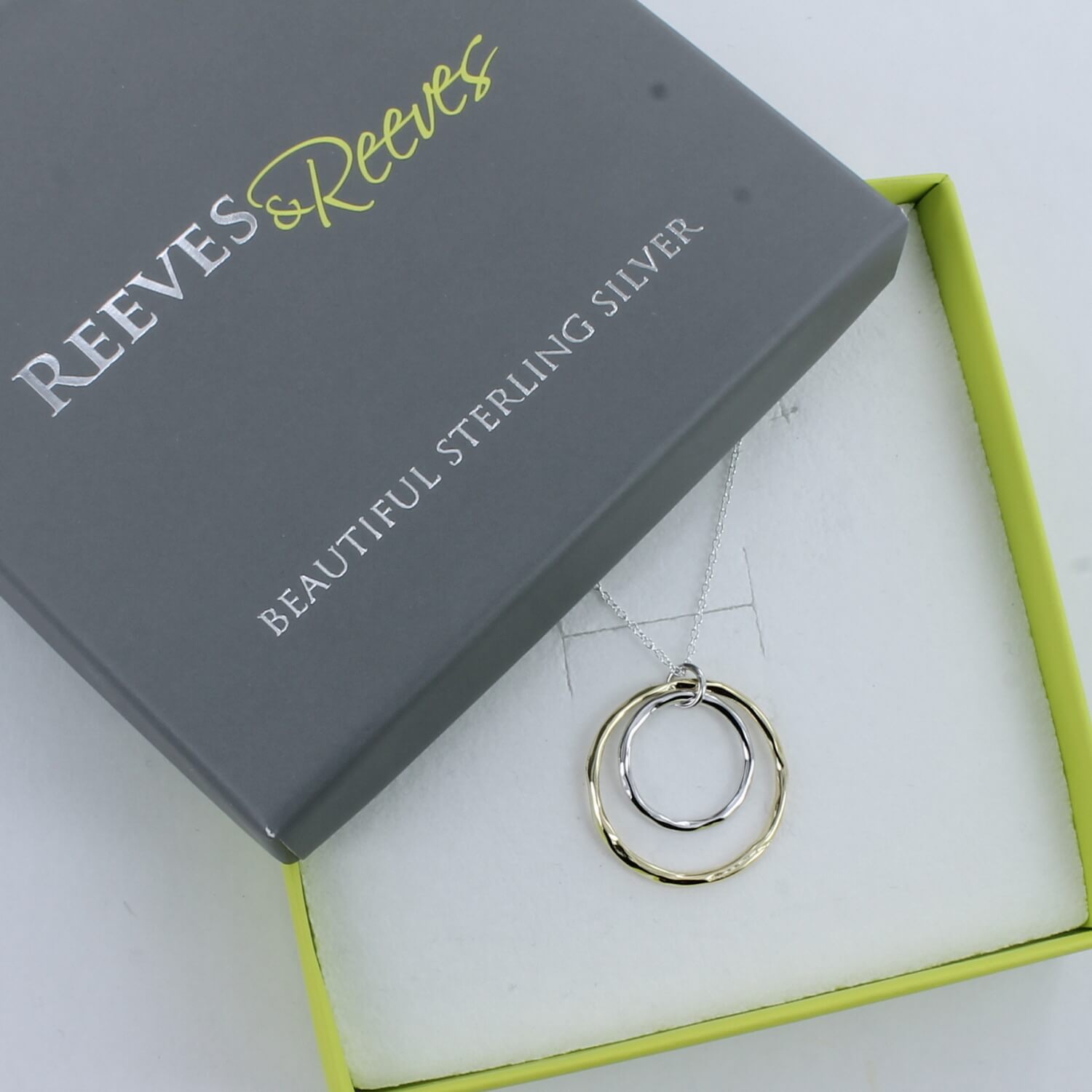 A gold pated circular pendant with a smaller silver circular pendant sat inside. Both hang from a silver chain and are shown in a Reeves & Reeves branded gift box.