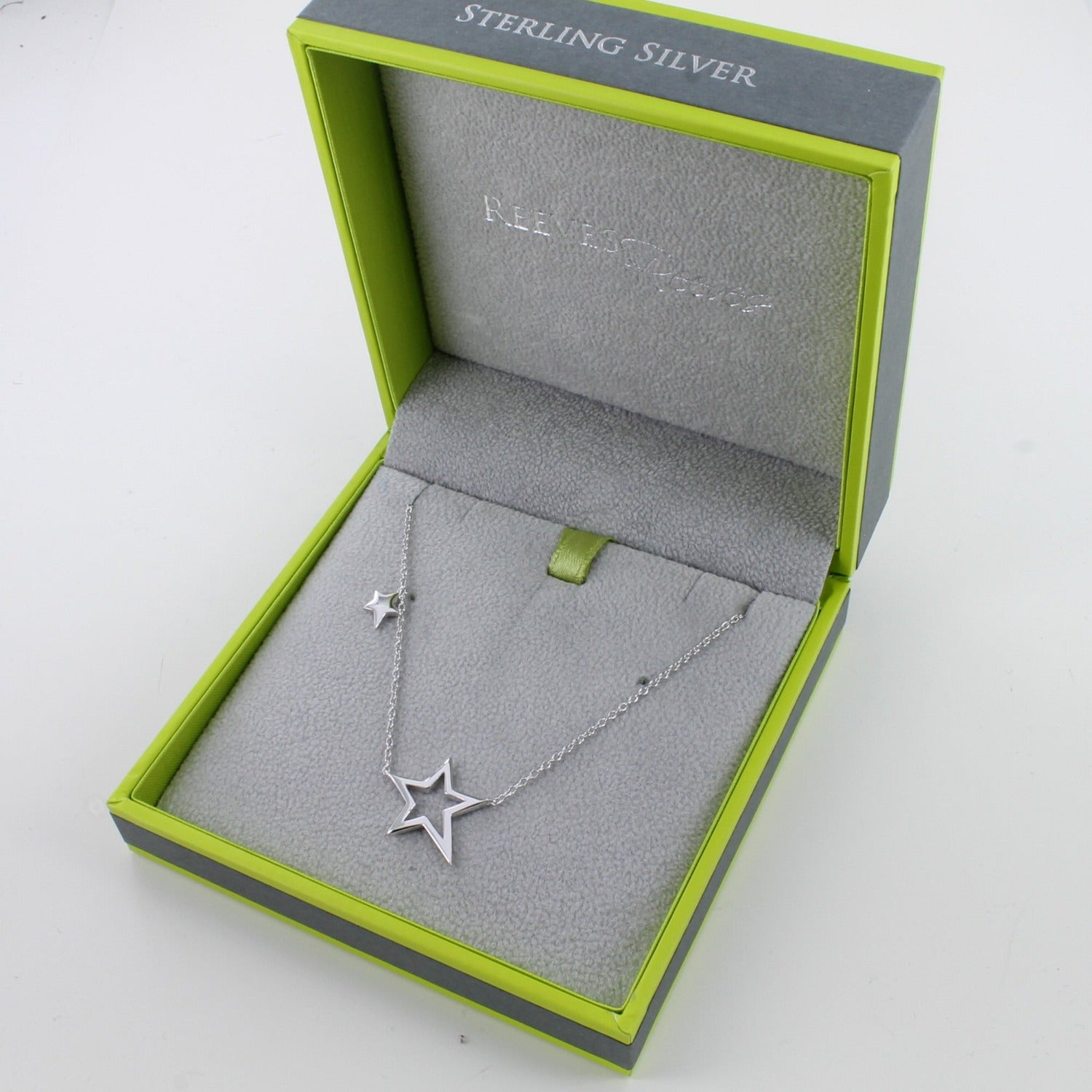 Silver necklace with a silhouette star shape at the centre and a smaller solid star further up the chain. Shown in a Reeves & Reeves branded gift box in green and grey colours.