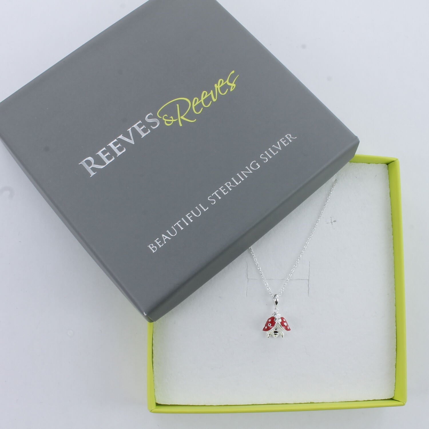 Sterling Silver Ladybird Necklace with red enamel detail on its wings and hung on silver chains. Shown in a Reeves & Reeves grey and green branded gift box.