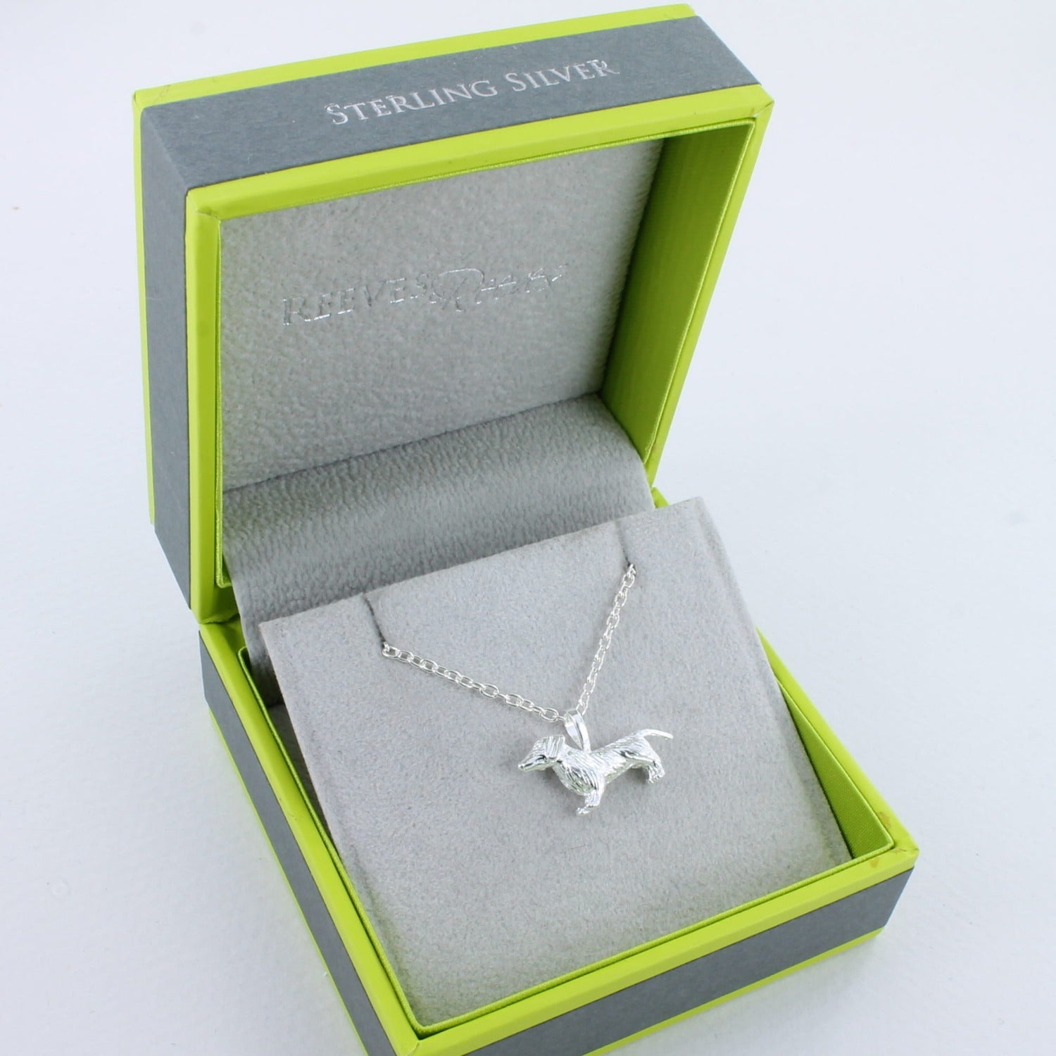 A silver dachshund dog pendant on a silver chain, shown in a Reeves & Reeves grey and green branded gift box.