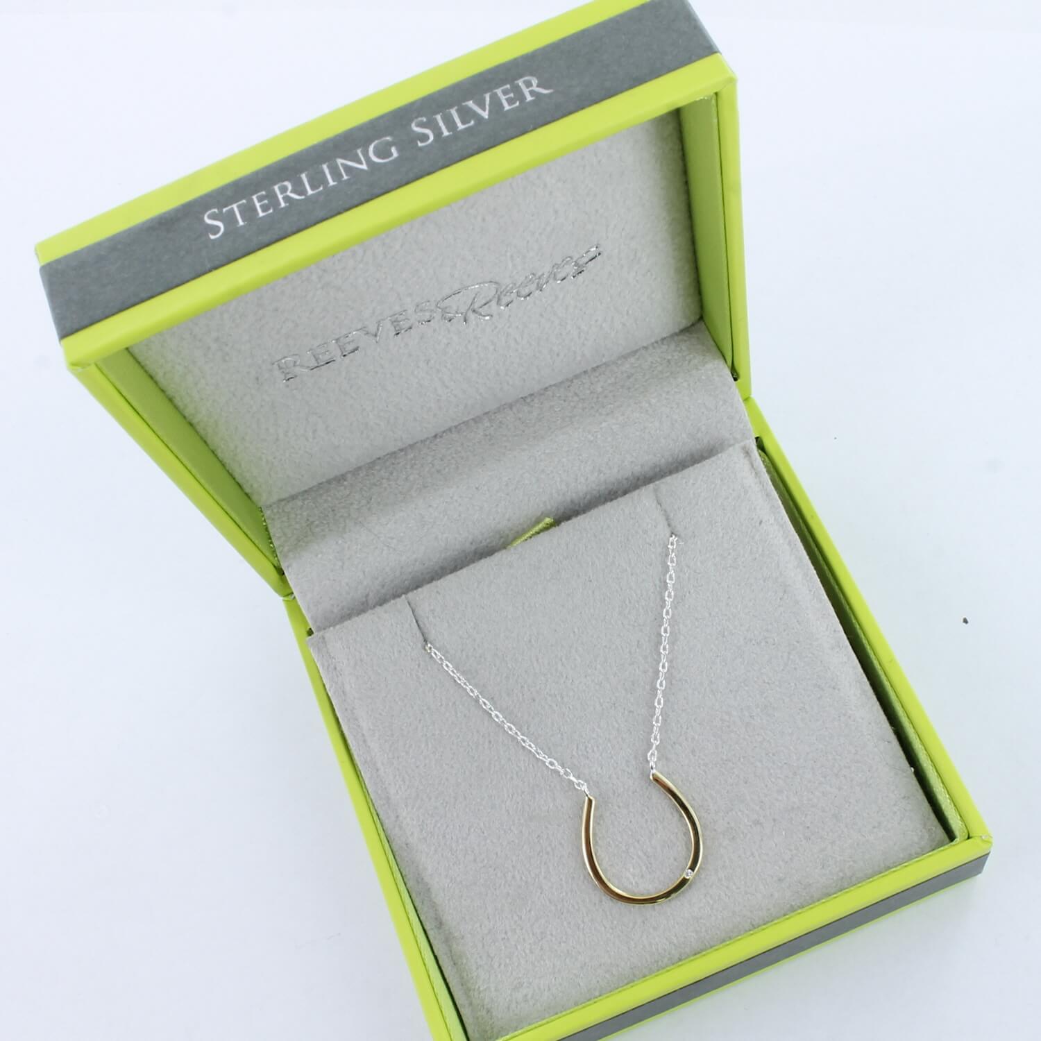 A silver and gold plated horseshoe necklace with a cubic zirconia stone, hung centrally on a fine silver chain. Shown in a Reeves & Reeves grey and green branded box.