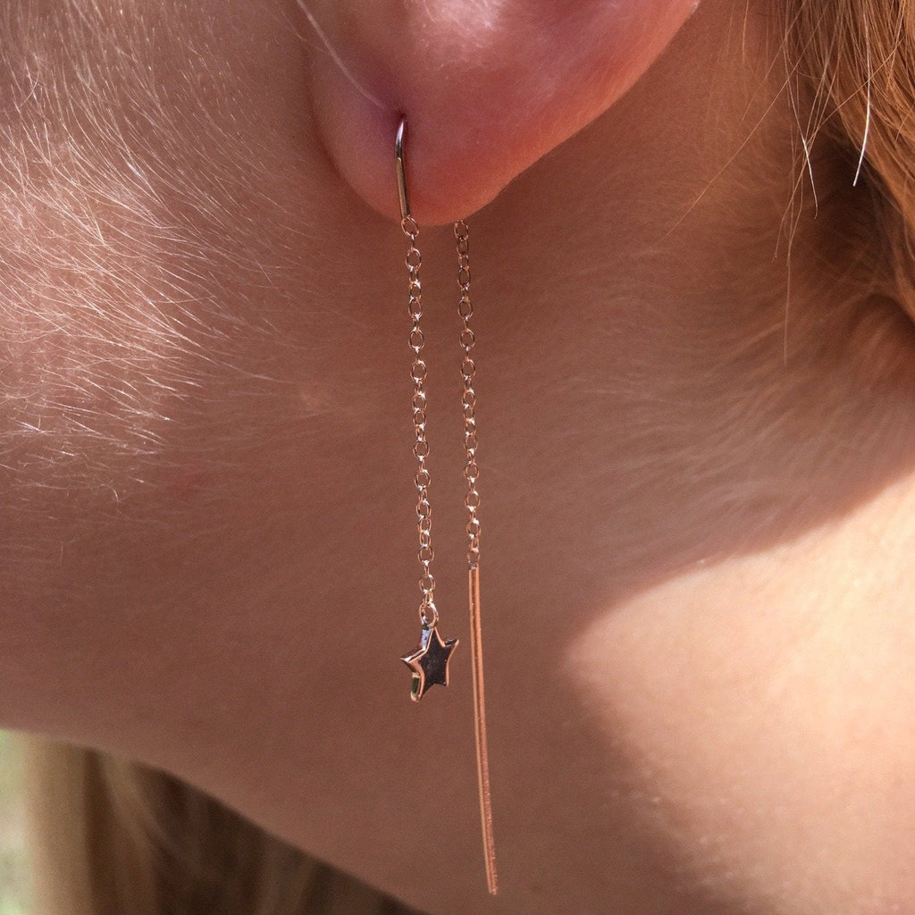 A pull through earring in sterling silver and 18ct rose gold plate, with a star charm at the bottom of a chain, threaded through a ladies ear with a charm and bar hanging behind the lobe.
