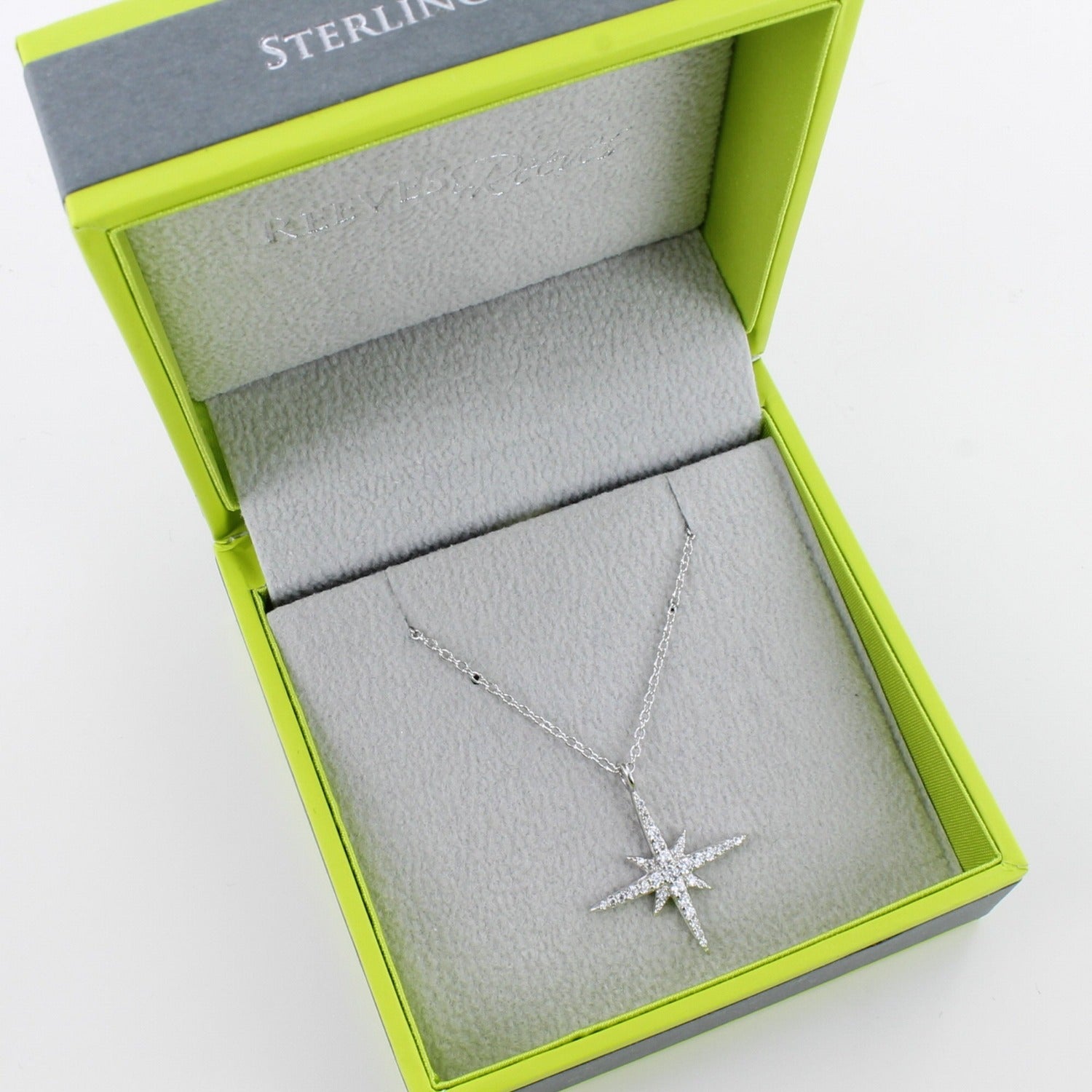 Sparkly Sterling Silver eight point star necklace, filled with cubic zirconia stones and hanging on a silver adjustable chain. Shown in a Reeves & Reeves branded grey and green gift box.