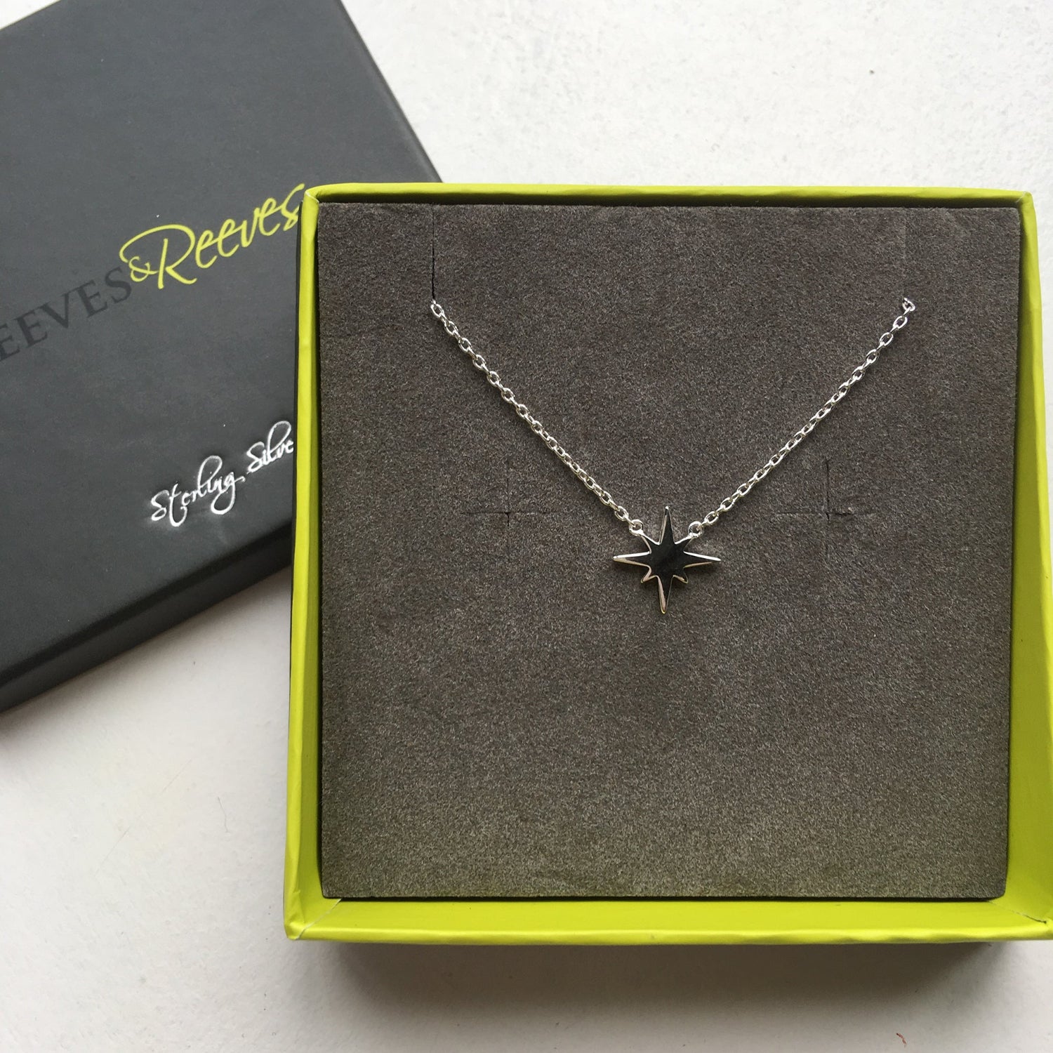 Sterling Silver eight point star necklace, sat centrally on a fine silver chain and shown in a Reeves & Reeves grey and green branded gift box.