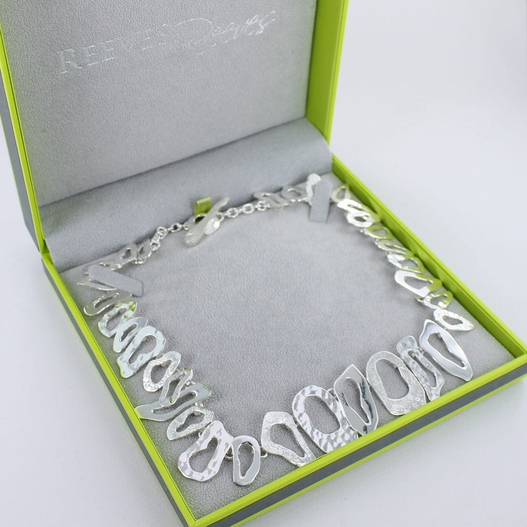 A sterling silver necklace with irregular shapes that have varying textures, shown in a Reeves & Reeves grey and green gift box.