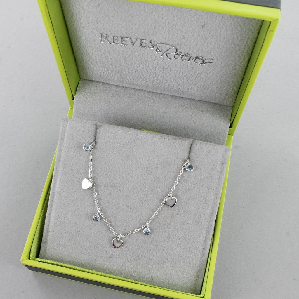A sterling silver chain necklace, with flat silver heart charms spread evenly along the chain, interspersed with blue gemstones. Shown in a Reeves & Reeves grey and green branded gift box.