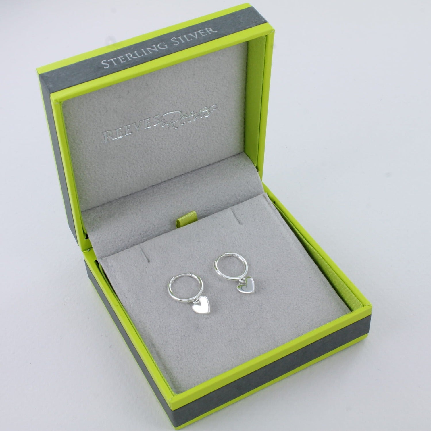A pair of sterling silver hoop style earrings with a silver heart charm hanging from the bottom of each hoop. Shown in a Reeves & Reeves grey and green gift box.
