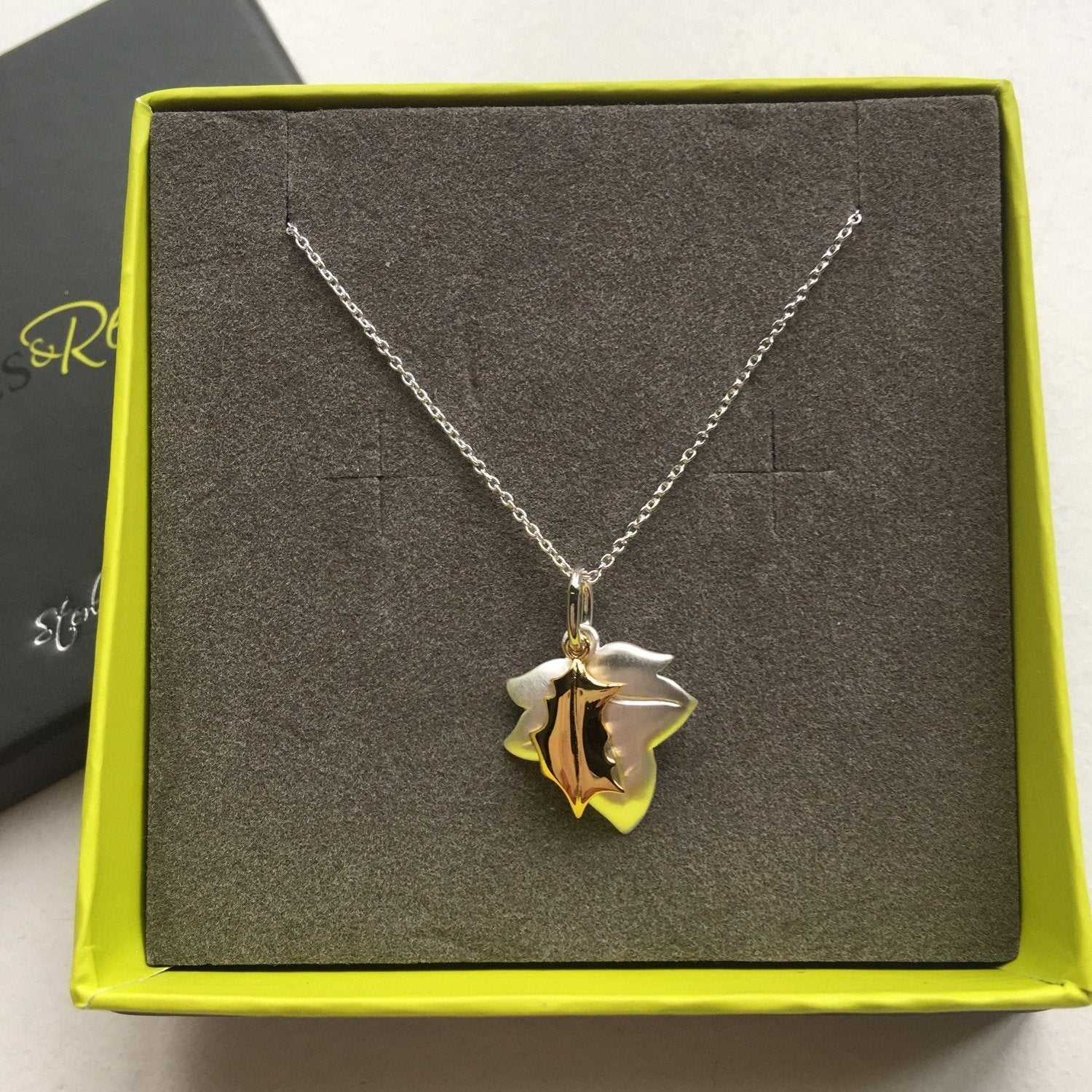 A silver ivy leaf charm with a smaller gold plated holly leaf charm hanging alongside, on a silver chain and shown in a Reeves & Reeves gift box.