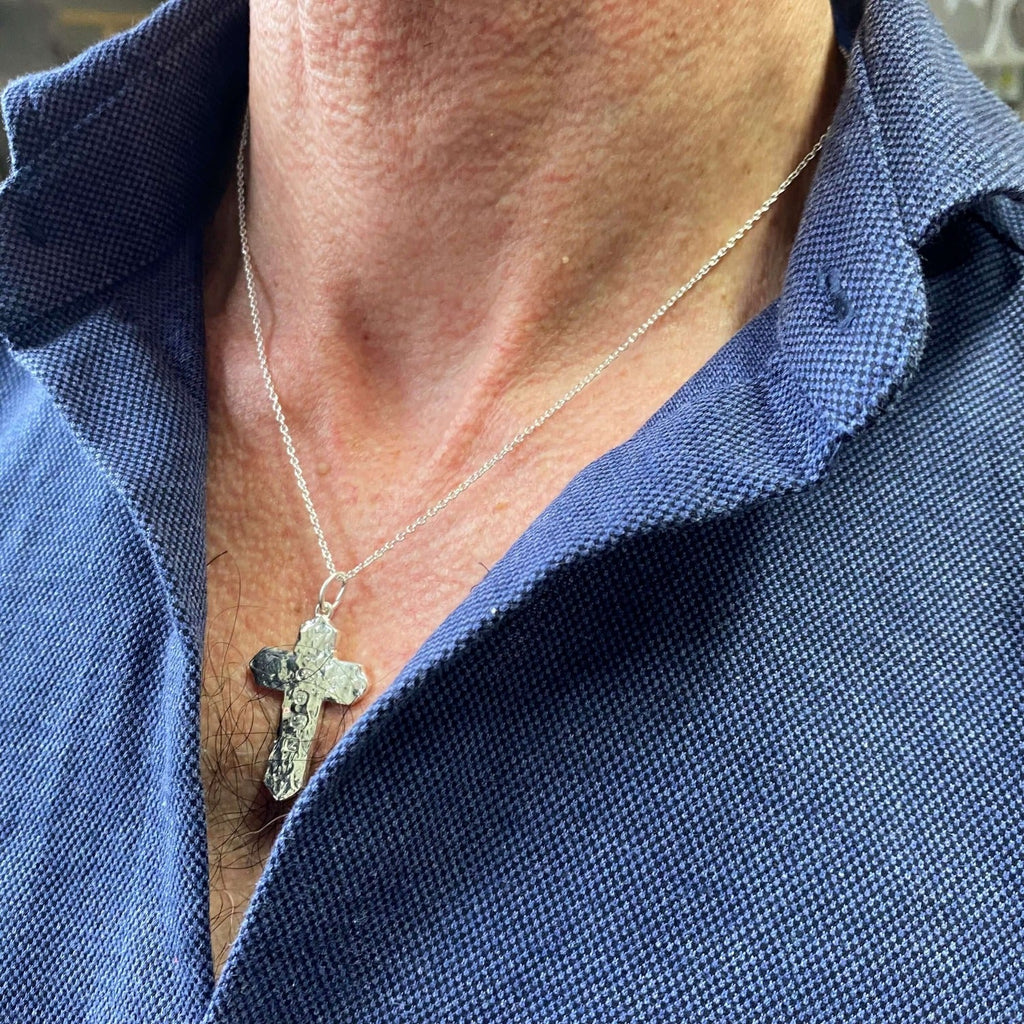 A sterling silver large cross pendant with hammered detail and hung on a silver chain. Worn on a man's neck.