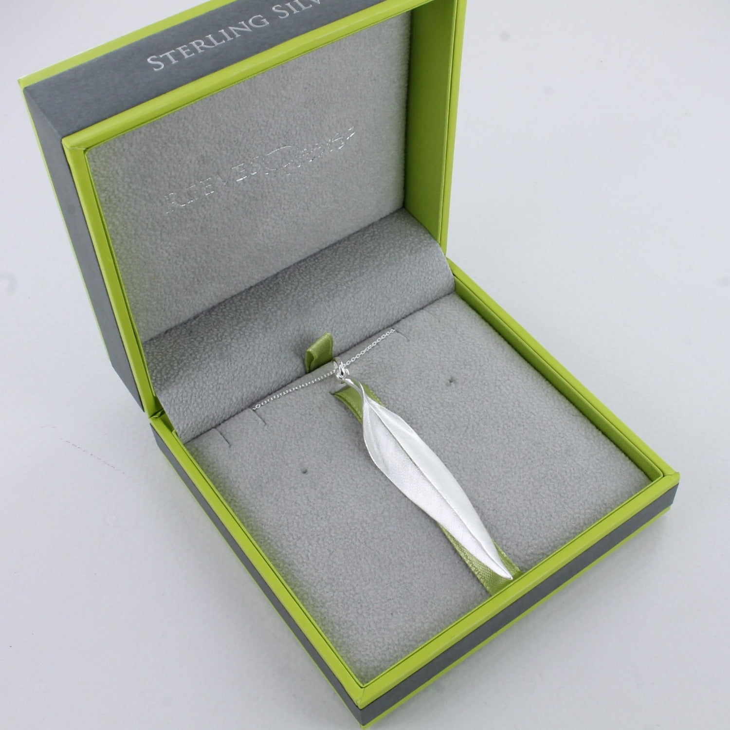 A Sterling Silver Leaf pendant which has a matt finish and gently curved edges. The leaf hangs on a silver chain and is shown in a grey and green Reeves & Reeves branded gift box.