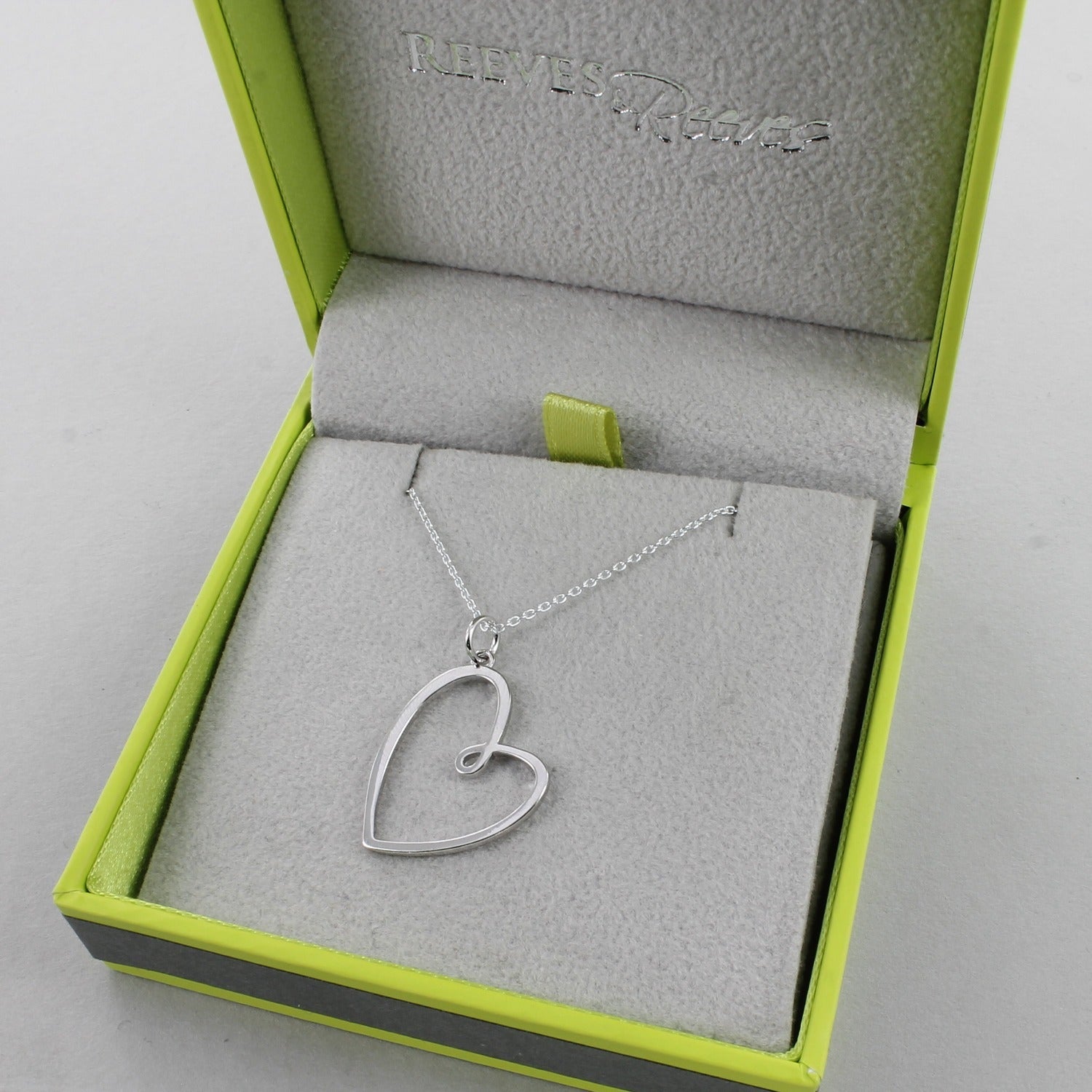A silver heart pendant hanging on a sterling silver adjustable chain. Shown in a Reeves & Reeves branded box.