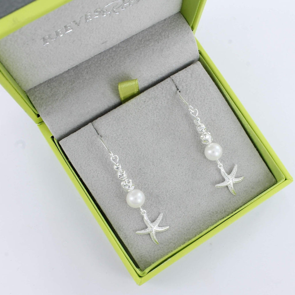 A pair of sterling silver drop style earrings with textured silver beads and a white freshwater pearl with a textured starfish hanging from the bottom. Shown in a Reeves & Reeves gift box.