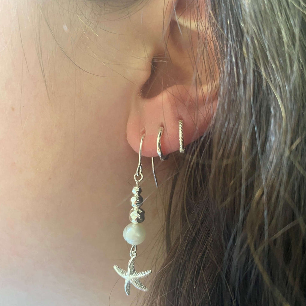 A starfish drop earring with three hammered silver beads hanging vertically with a white freshwater pearl at the bottom. From the bottom a textured starfish hangs. Shown in a ladies ear.