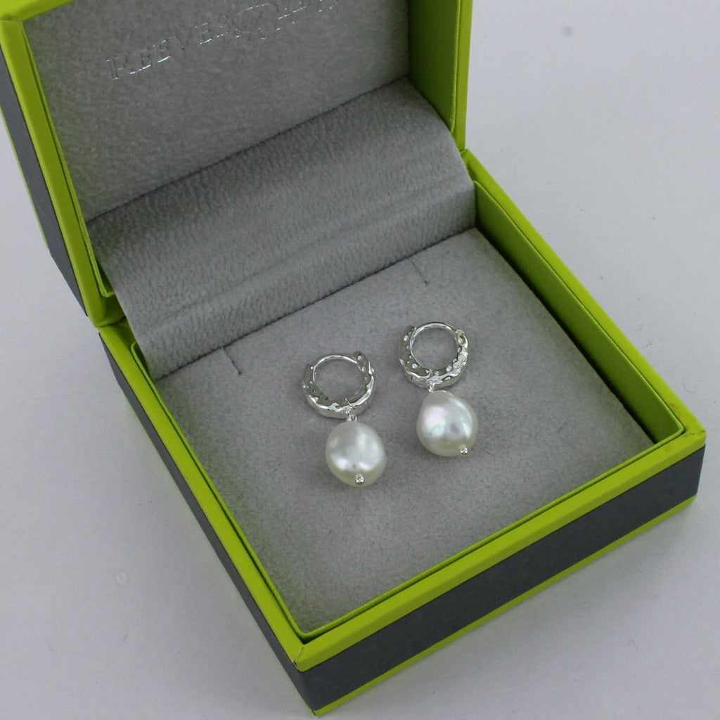 A sterling silver pair of textured hoop earrings with a white freshwater pearl hanging from the bottom. Shown in a Reeves & Reeves gift box.
