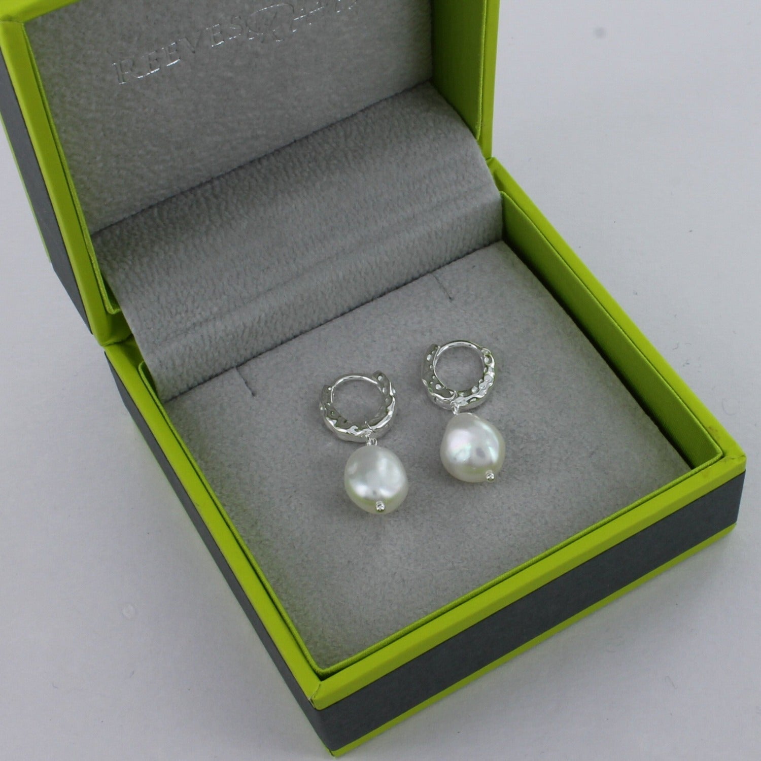 A sterling silver pair of textured hoop earrings with a white freshwater pearl hanging from the bottom. Shown in a Reeves & Reeves gift box.