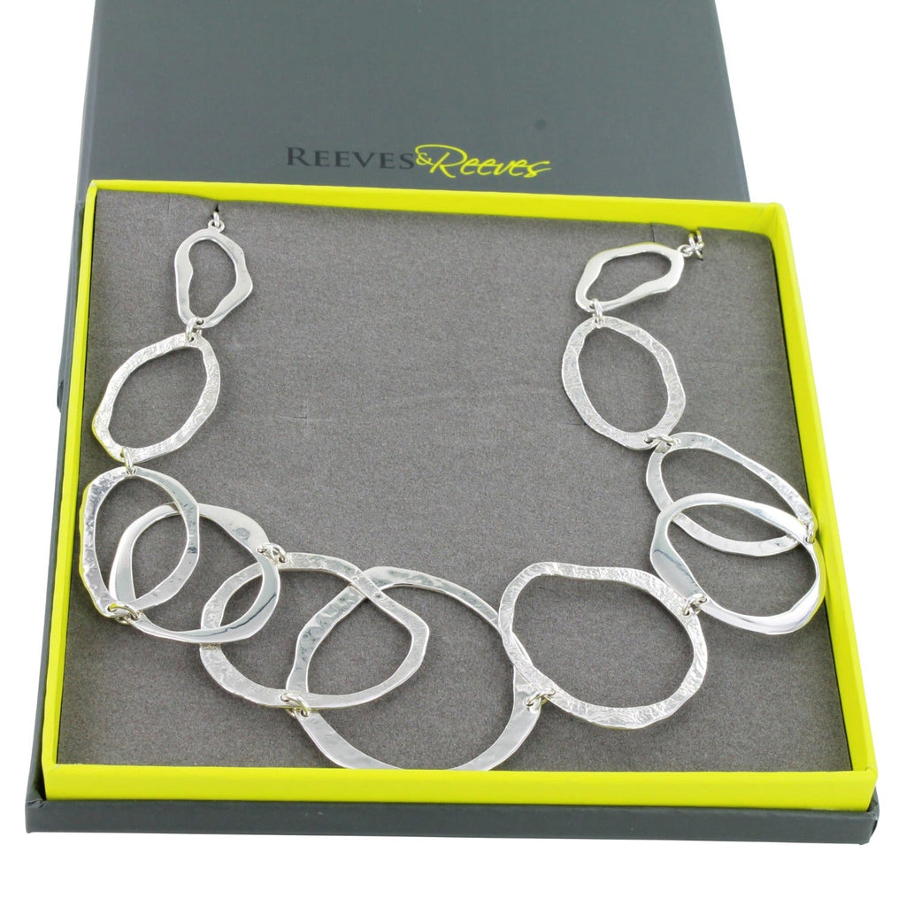 A sterling silver circular outline necklace, all the circles have varying textures and the necklace is shown in a Reeves & Reeves grey and green branded gift box.