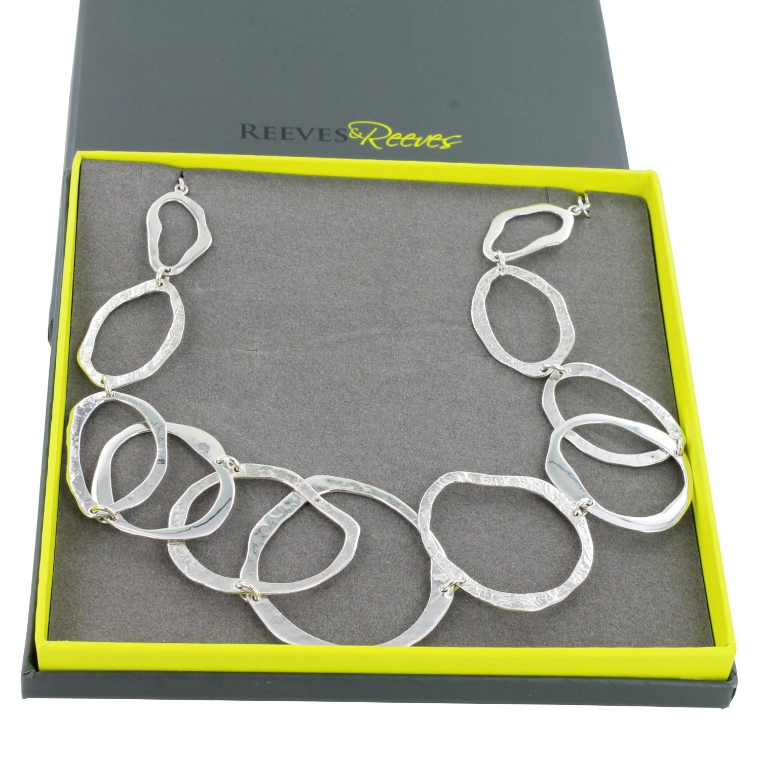 A sterling silver circular outline necklace, all the circles have varying textures and the necklace is shown in a Reeves & Reeves grey and green branded gift box.