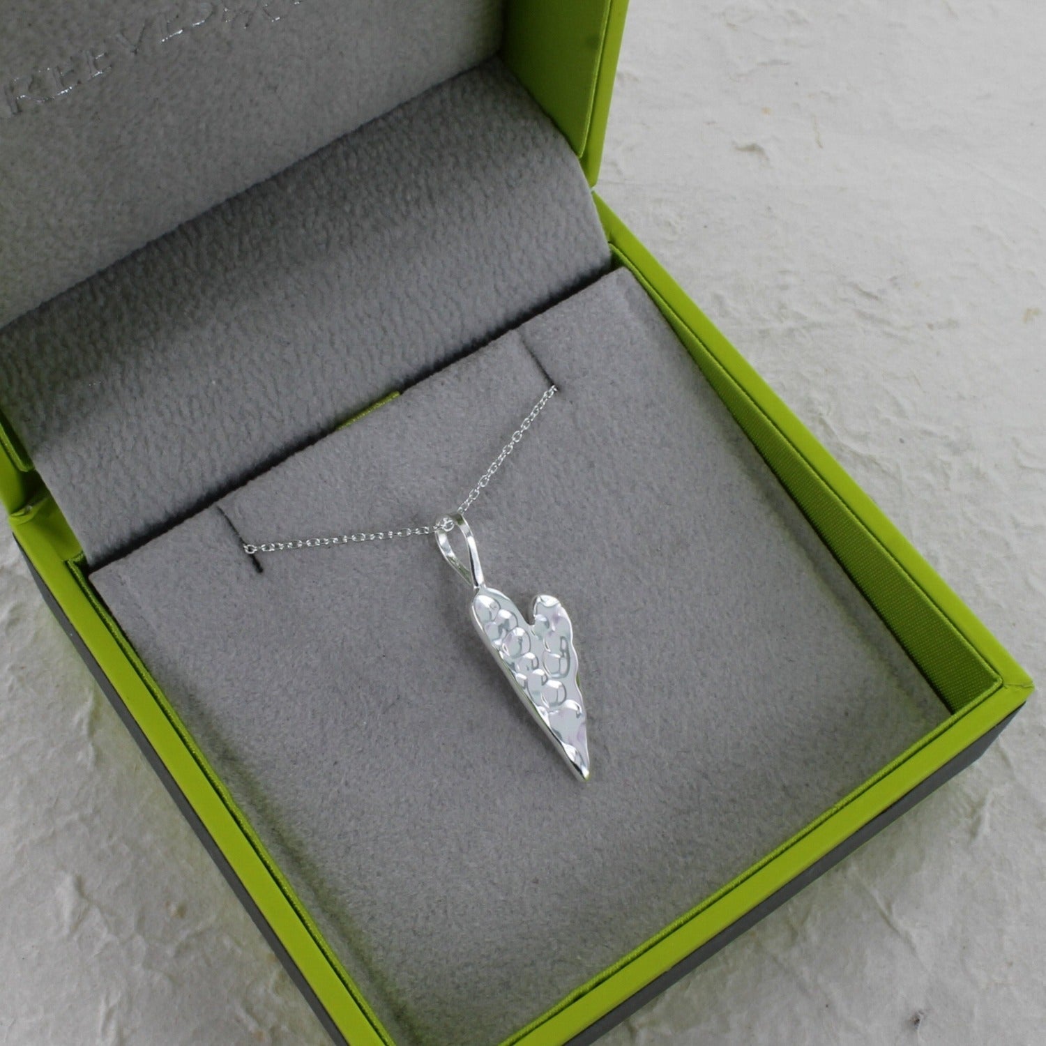A silver textured heart pendant on an adjustable silver chain and shown in a Reeves & Reeves grey and green gift box.