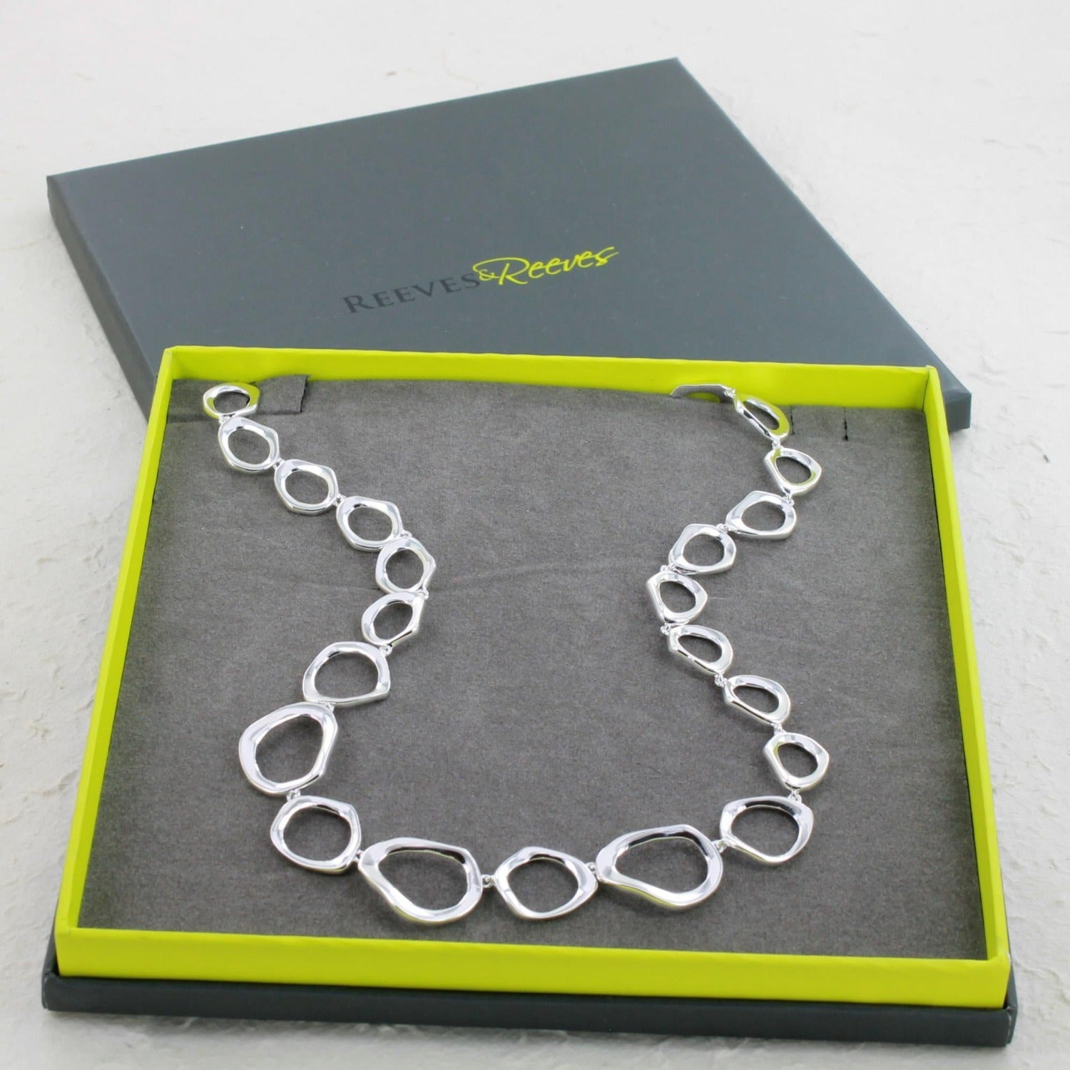 A sterling silver ringlets necklace, comprised of rings with undulated edges, in a continuous line. Shown in a Reeves & Reeves grey and green branded gift box.