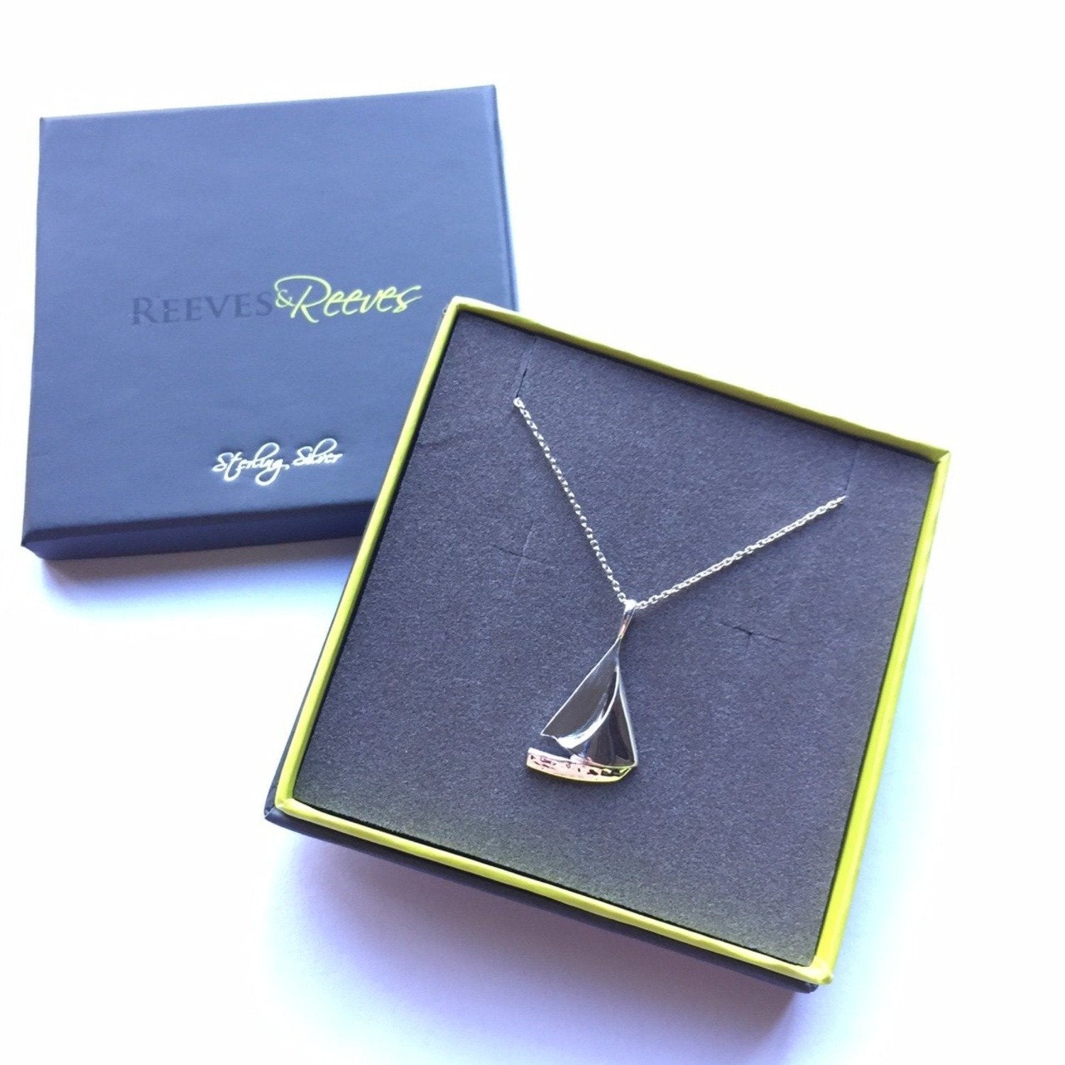 A silver sailboat pendant on a silver chain and shown in a Reeves & Reeves branded gift box.
