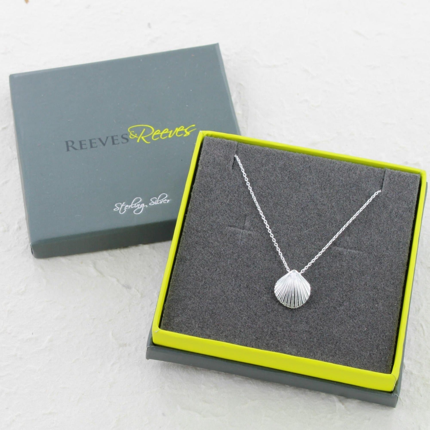 A silver scallop shell necklace on a silver chain and shown in a Reeves & Reeves grey and green branded gift box.