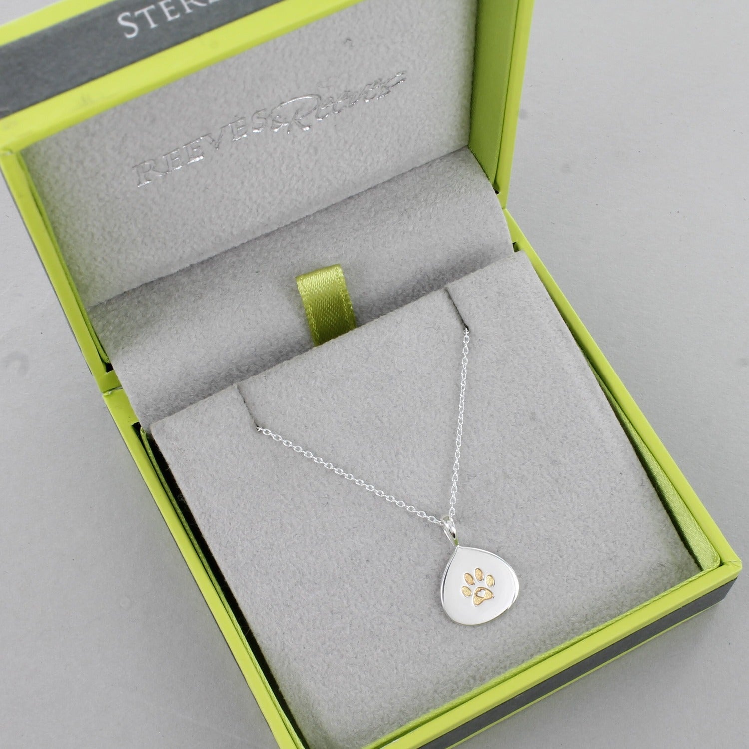 A silver teardrop pendant with a gold plated pawprint design in the centre. Hangs on a silver adjustable chain and shown in a Reeves & Reeves gift box.