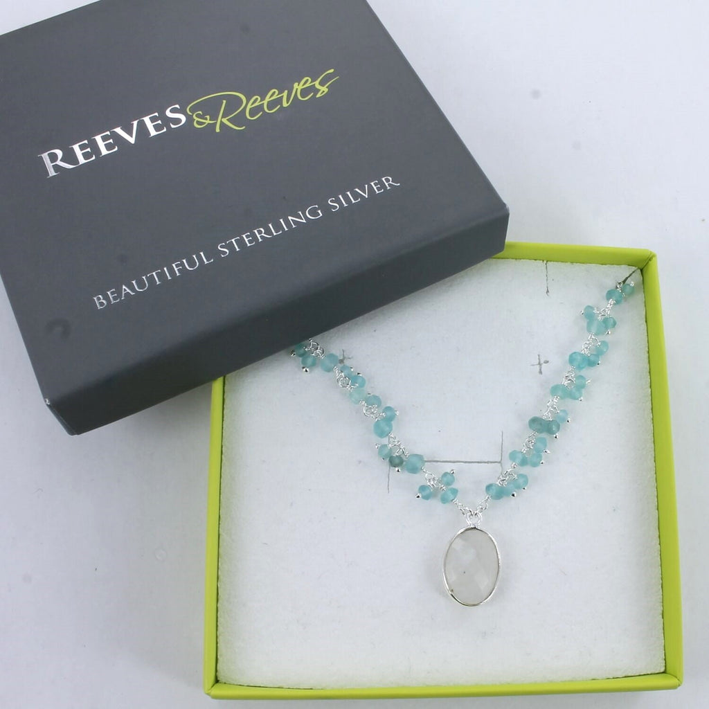 A silver chain necklace with an oval shaped moonstone hanging from the centre of the chain. Small Aqua chalcedony stones sit in clusters up either side of the chain. Shown in a Reeves & Reeves gift box.