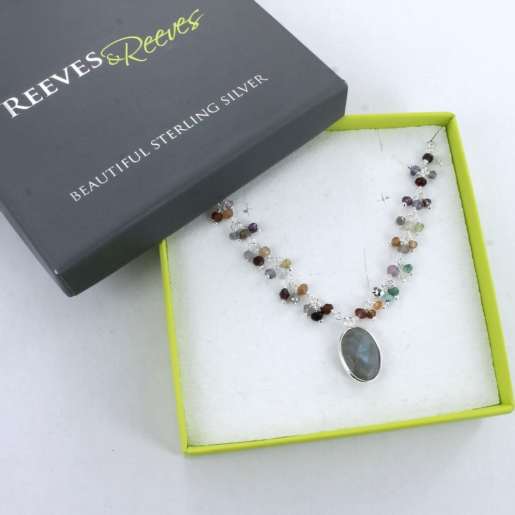 A sterling silver fine chain necklace with an oval labradorite stone set at the centre and more smaller coloured stones sit up either side of the chain. Shown in a Reeves & Reeves gift box.