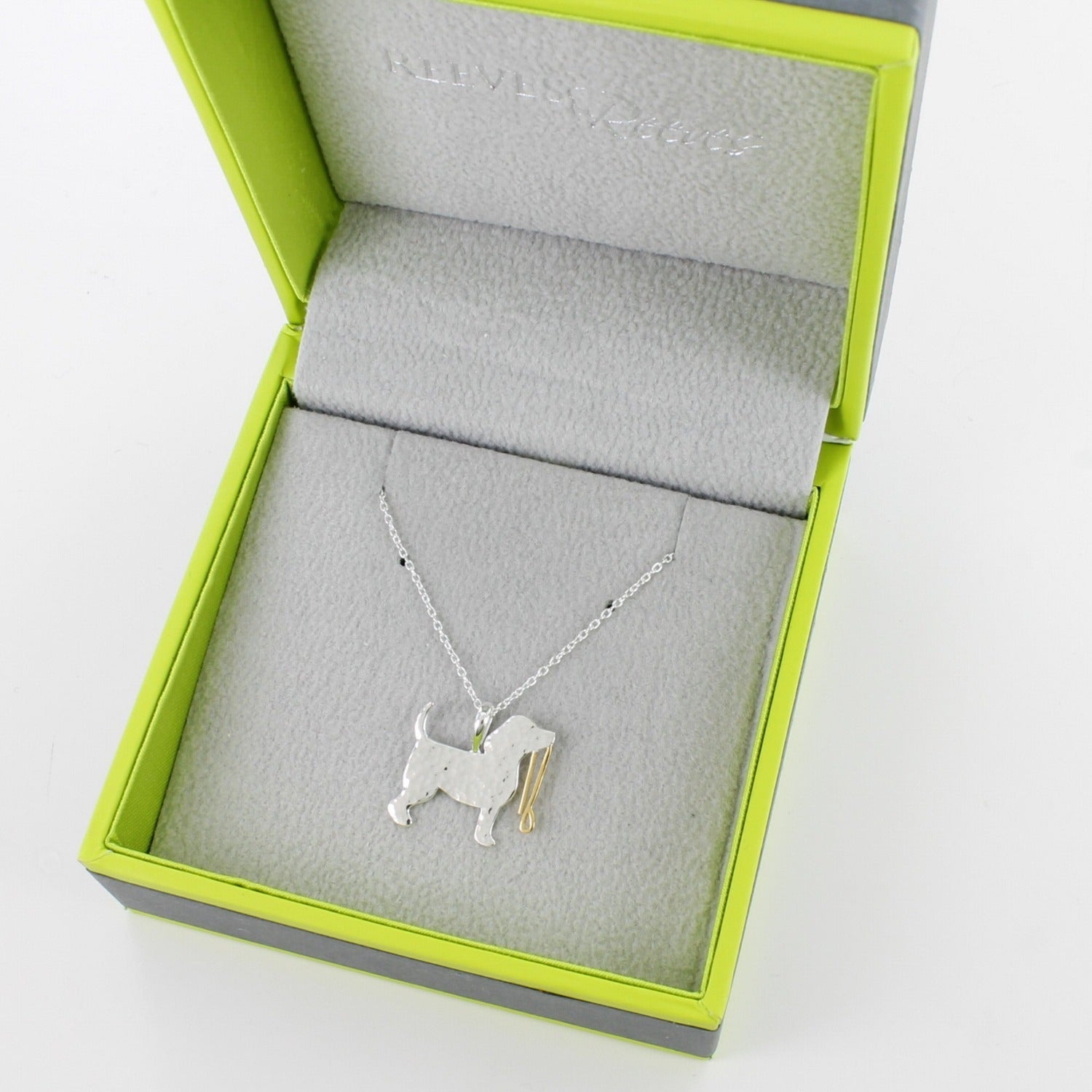 A silver dog pendant with a hammered finish and holding a gold plated lead in its mouth, hanging on a silver chain. Shown in a Reeves & Reeves branded gift box.