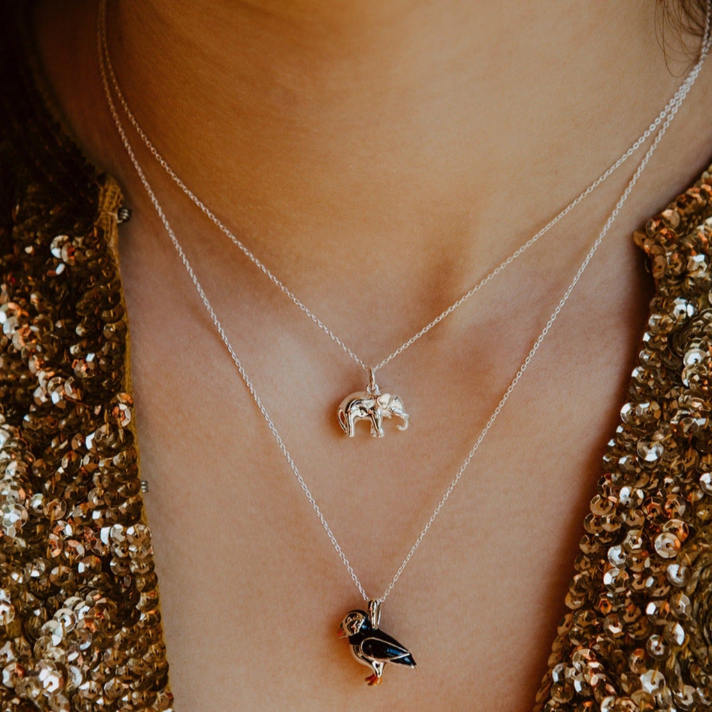 A sterling silver 3D elephant pendant, hanging on an adjustable chain and worn on a neck. Paired with an enamel black and orange puffin necklace.