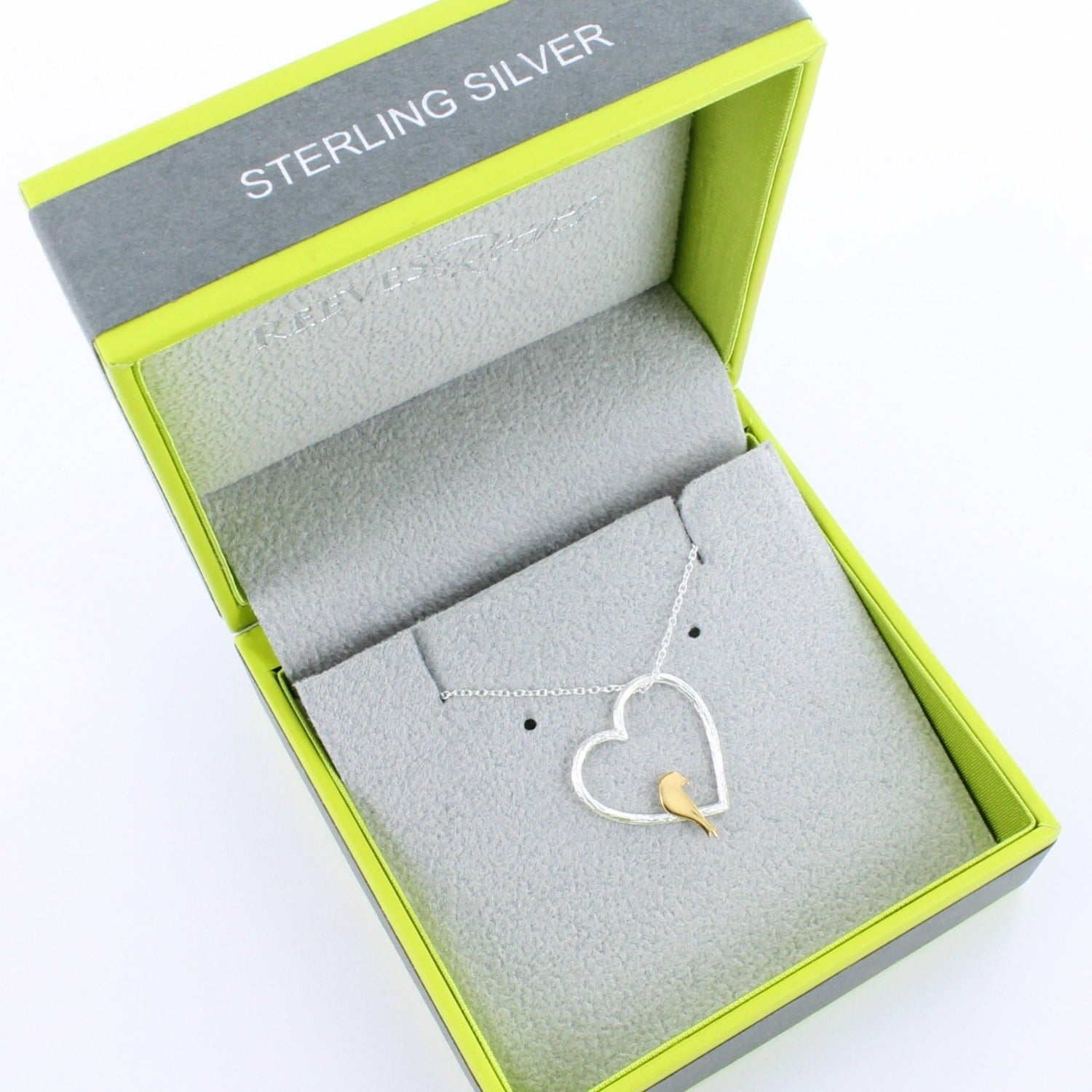 Silver silhouette of a heart with an 18ct gold plate bird perched on the edge. The heart pendant sits on an adjustable silver chain and is shown inside a Reeves & Reeves grey & green gift box.