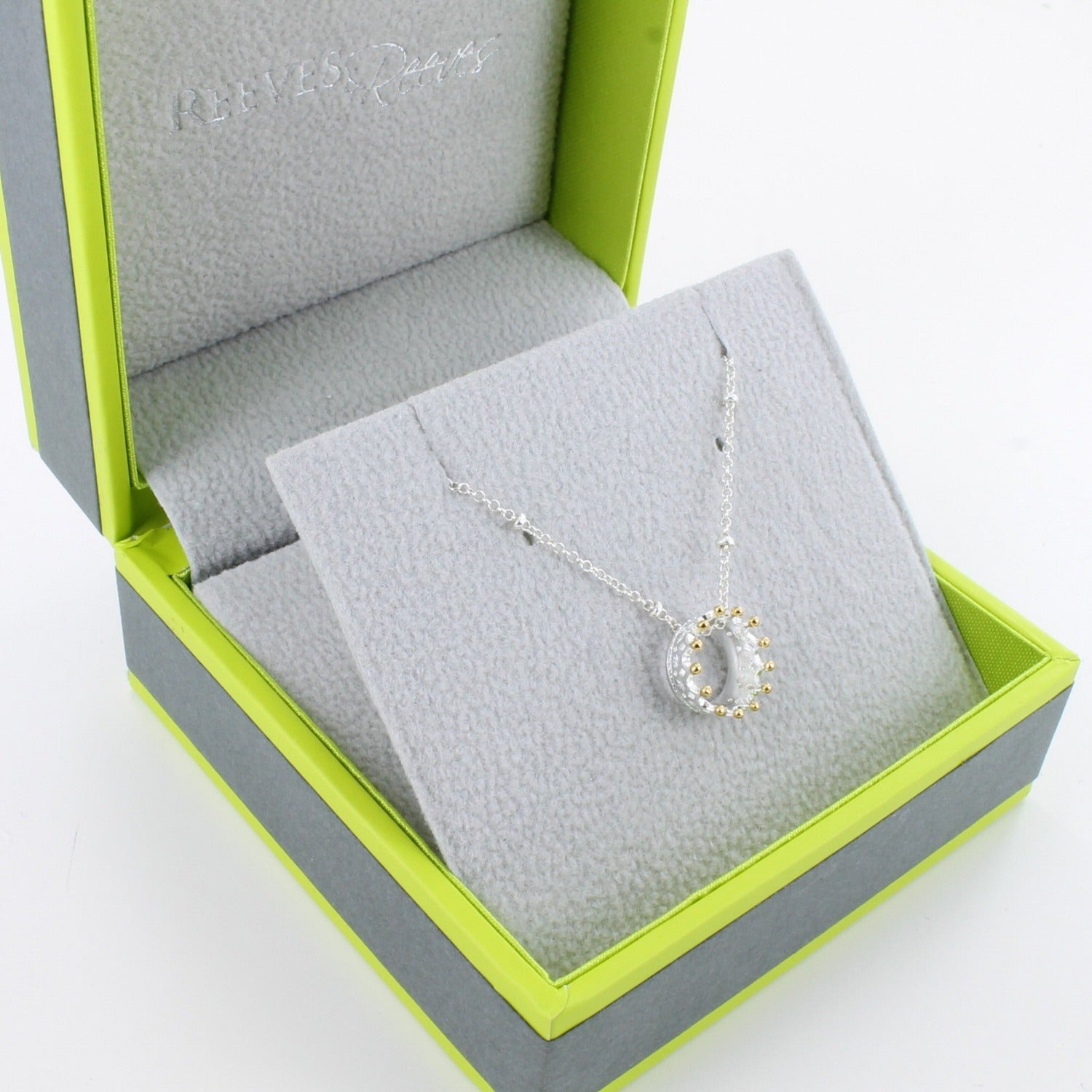 A silver crown necklace with gold plated finish on its spikes, hung on a silver beaded chain and shown in a Reeves & Reeves branded gift box.