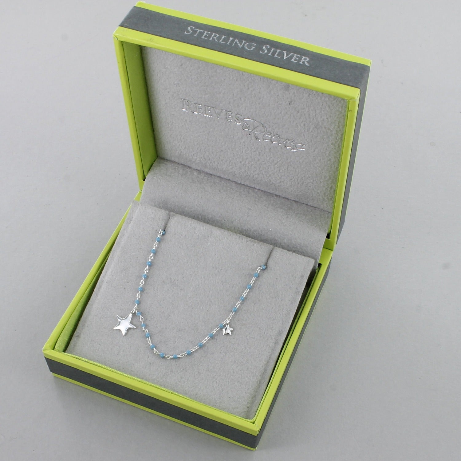 A sterling silver and baby blue enamel chain necklace, with a silver star at the centre and a smaller star further up the chain. Shown in a grey and green Reeves & Reeves gift box.
