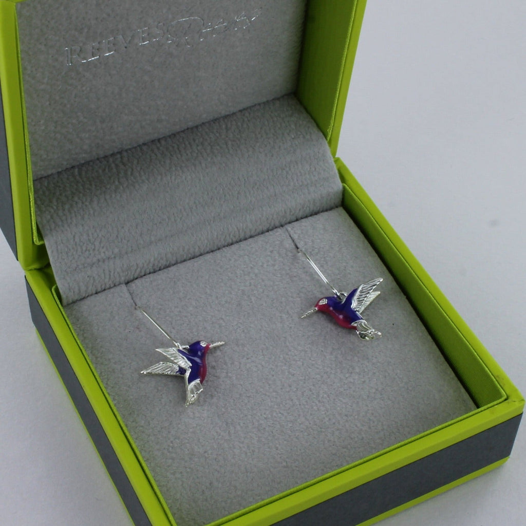A sterling silver pair of hummingbird earrings with pink and purple enamel detail, hanging on silver hooks. Shown in a Reeves & Reeves grey and green gift box.